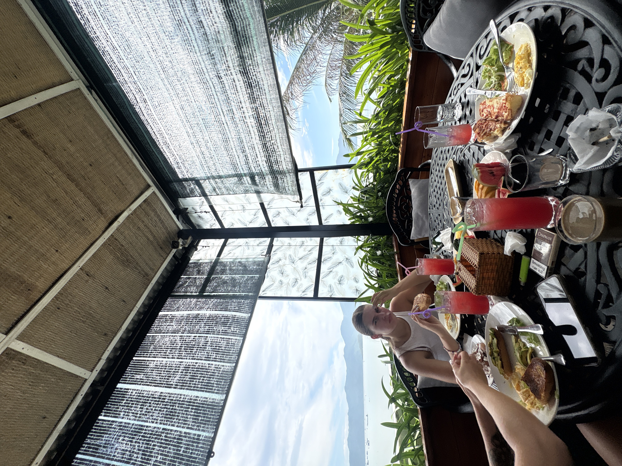 The image depicts a group of individuals seated at an outdoor table, enjoying a meal and drinks. The table is laden with plates of food, glasses filled with red beverages, and a basket. The individuals appear to be engaged in conversation and dining. The setting suggests a restaurant or cafe with a view of the ocean and mountains in the background. The architecture includes a thatched roof and decorative screens. The overall ambiance is relaxed and casual, indicative of a tourist destination. Based on the provided context of Bắc Nha Trang, Vietnam, this scene likely takes place at a waterfront establishment offering scenic views.