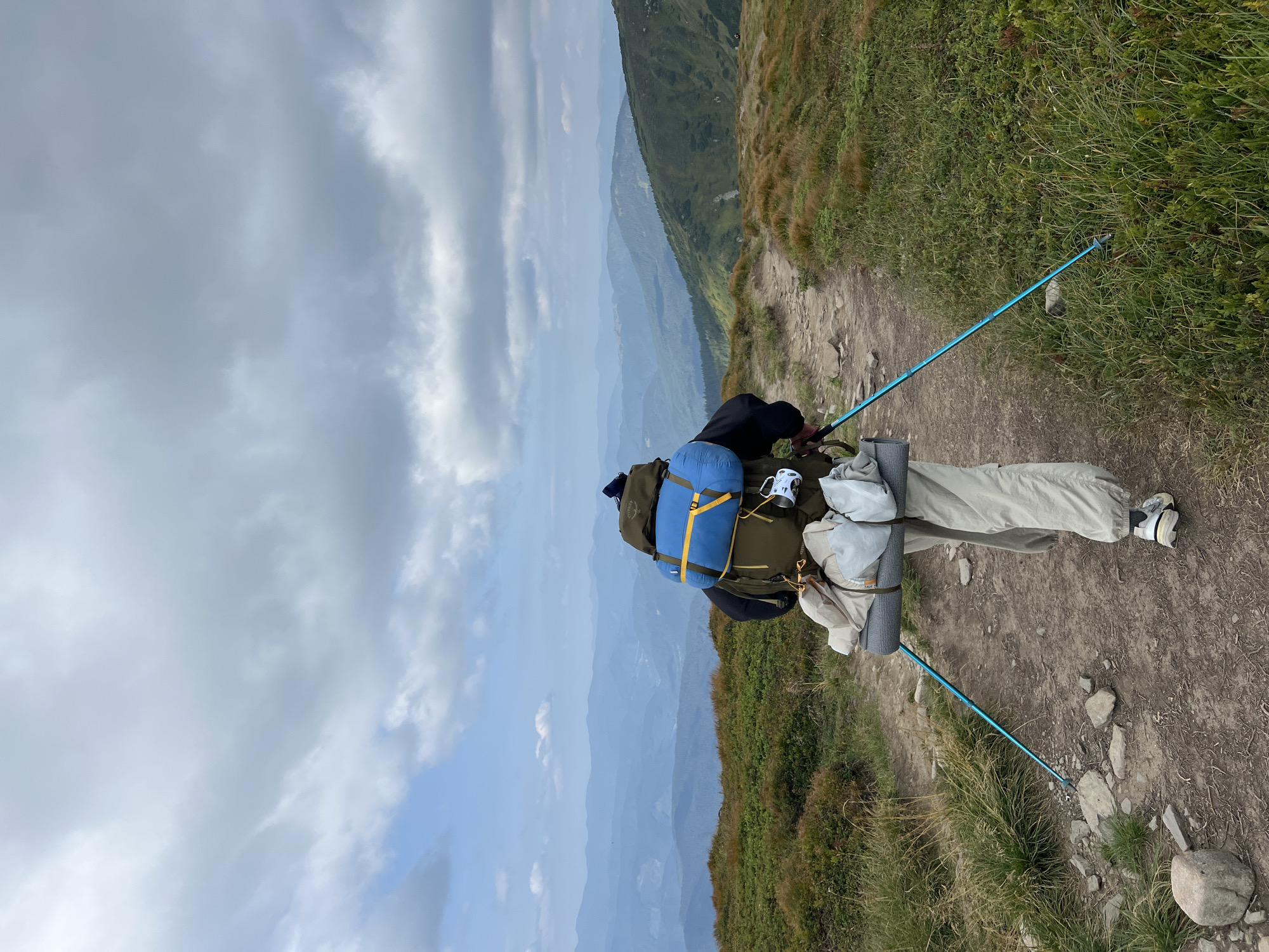 In this vertically oriented photograph, a lone hiker is captured from behind, traversing a dirt path in a mountainous terrain. The hiker is equipped with a large backpack containing a blue sleeping bag, a rolled sleeping pad secured to the side, and other items. They are also using two trekking poles, one of which is extended forward, to aid their movement. The path itself is rough and stony, winding uphill through grassy slopes.
The surrounding environment is dominated by rolling hills and distant mountain ranges, layered in shades of blue and green, suggesting a vast expanse. The vegetation consists of low-lying grasses and shrubs, with some patches of brown indicating drier areas. The sky above is partly cloudy, with a mix of white and gray clouds against a pale blue backdrop. The scene evokes a sense of solitude and adventure, typical of a hiking expedition in a natural, elevated landscape. The location is identified as the city of Hoverla, Ukraine, which is known for its mountainous geography and is home to Mount Hoverla, the highest peak in Ukraine.