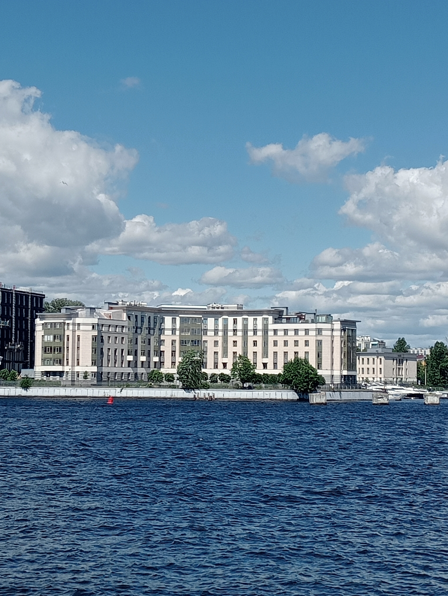 This media file depicts a scenic view of a waterfront on a bright, partly cloudy day in Saint Petersburg, Russia. The foreground is dominated by the deep blue, choppy waters of a river or canal, reflecting the sky. A red buoy is visible in the water, marking a navigable channel.
The midground features a modern, multi-story building complex with a light-colored facade, characterized by numerous windows and balconies. Adjacent to this main building, other structures of similar architectural style are visible, suggesting a developed urban area. Lush green trees and manicured landscaping line the waterfront, contrasting with the concrete embankment. Several white boats are moored or docked near the shore, hinting at leisurely activities or transportation.
The background is filled with a bright blue sky punctuated by scattered, fluffy white clouds. A faint silhouette of a bird can be seen in the upper left portion of the sky, adding a touch of life to the expansive heavens. The lighting suggests it is daytime, likely midday or early afternoon, given the brightness and the clarity of the shadows. The overall atmosphere is serene and picturesque, showcasing a modern riverside development within the historic city of Saint Petersburg. There is no visible text in the image.