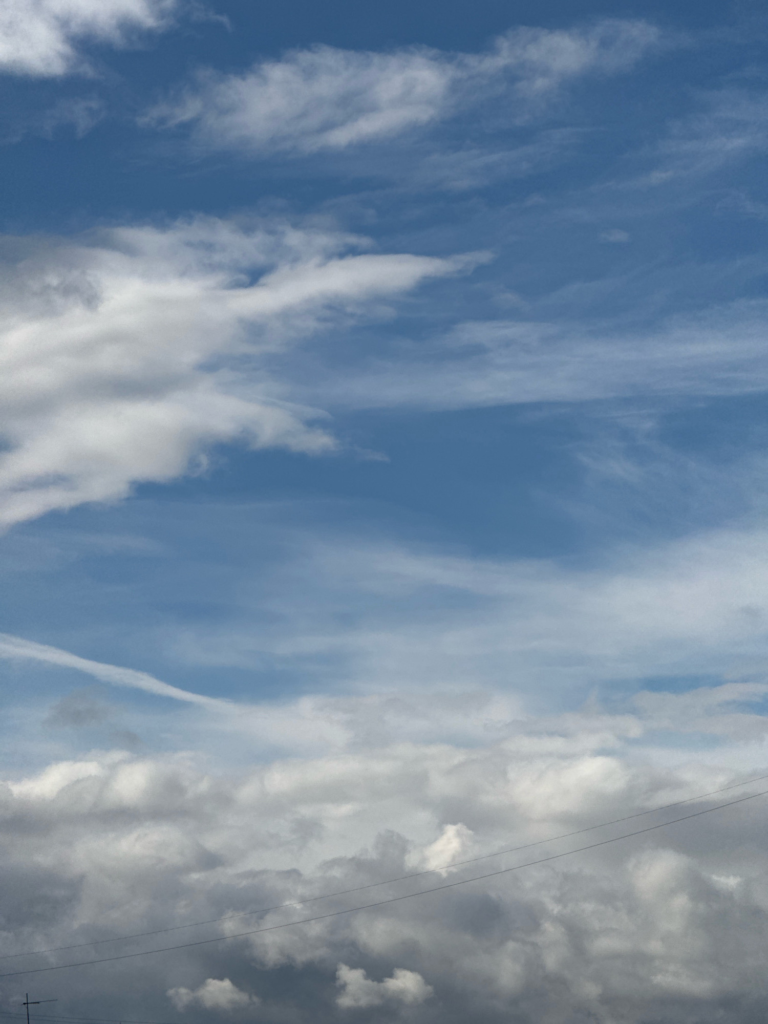 The image depicts a cloudy sky with a blue hue. The clouds are varied in texture and density, with some appearing wispy and others more substantial. A thin, linear object, possibly a power line, runs horizontally across the lower portion of the image. In the far lower-left corner, a portion of a cross-shaped structure is visible, suggestive of a utility pole or antenna. There are no people or discernible subjects or actions within the frame. The environment is solely characterized by the sky and the man-made linear elements. The location is identified as Krasnoyarsk, Russia, suggesting an outdoor scene within that city.