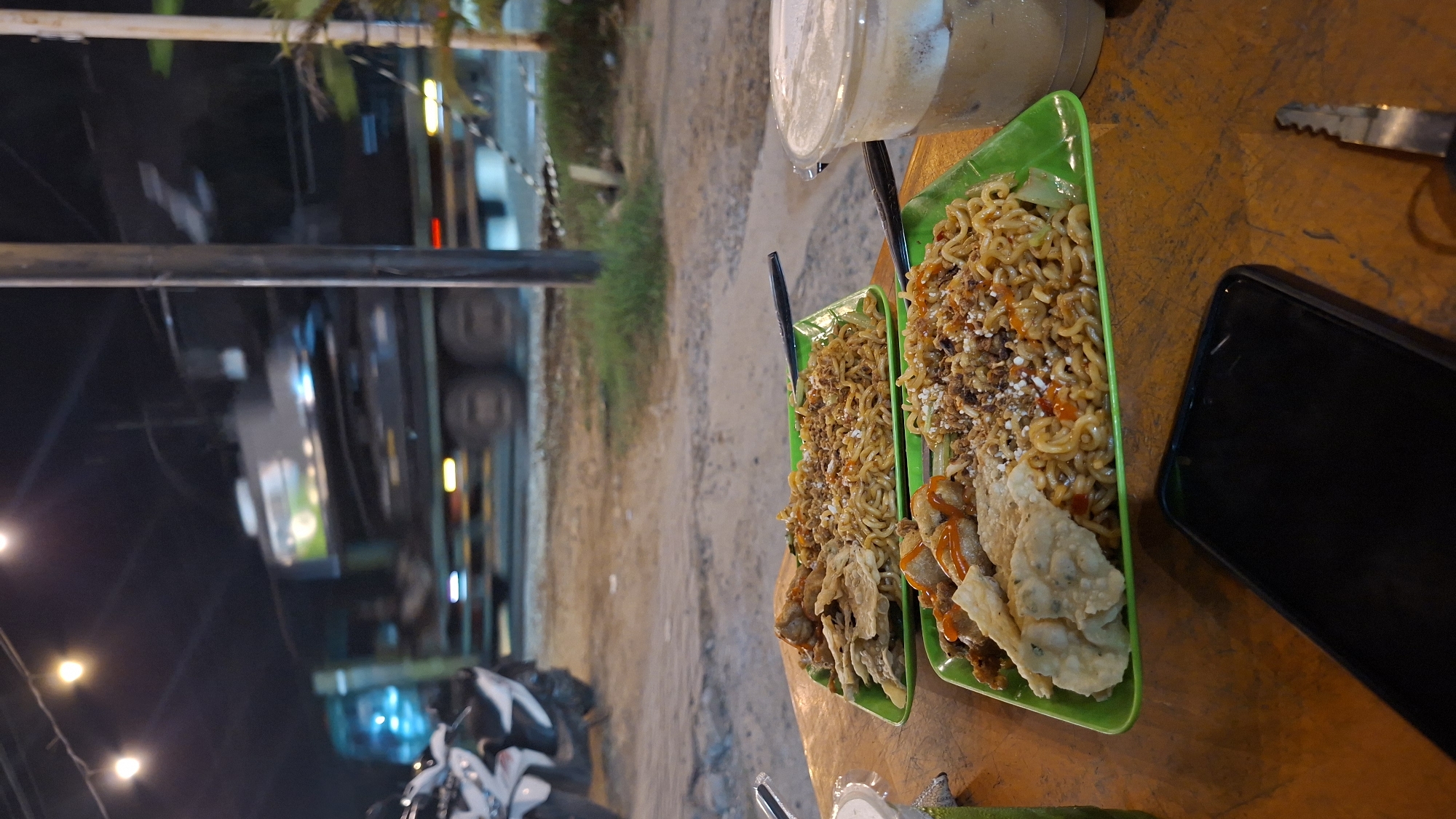 The image shows a dimly lit outdoor scene at night, likely a street food stall or casual dining area. In the foreground, two green rectangular plates of food are set on a wooden table. The plates contain noodles topped with various ingredients, including what appears to be fried dumplings or tempeh, and some are drizzled with sauce. A plastic cup filled with a layered iced beverage is also on the table to the right. In the lower right corner, a black smartphone and a car key are visible on the table.
In the background, a large truck with its lights illuminated is in motion on a road. A white motorcycle is parked to the left of the frame. The environment suggests a roadside setting with dirt or gravel on the ground and sparse vegetation. The overall impression is one of a humble, possibly late-night, dining experience in a public space. The mention of Keudeteupinpunti, Indonesia, places the scene within a specific geographical context.