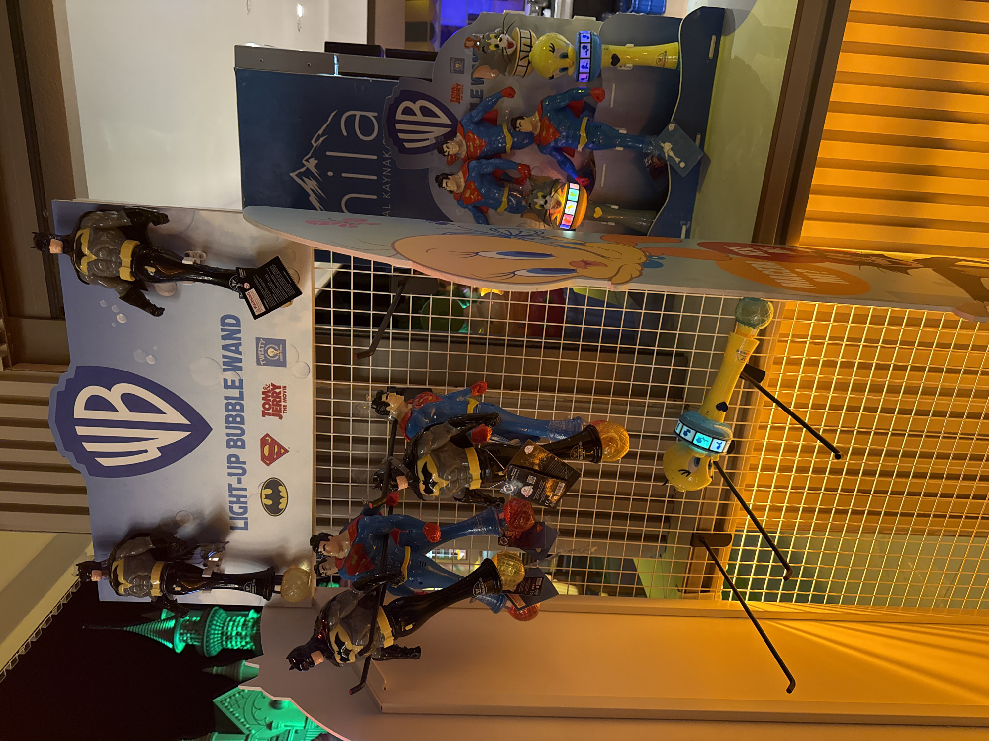 This image shows a retail display of Warner Bros. merchandise, specifically light-up bubble wands featuring Batman, Superman, Tom and Jerry, and Tweety Bird. The display is set up on a wire grid with several items hanging from hooks. On the left, two Batman bubble wands are prominently displayed. Behind them, two Superman bubble wands are visible. To the right, a larger display stand features multiple Superman bubble wands, a Tom and Jerry item, and Tweety Bird bubble wands. The background includes a large Warner Bros. logo and branding for "Ahila Al Kaynak" in Bodrum, Turkey. The lighting is a warm, yellow hue, casting shadows on the display. In the far left background, a green-lit tower can be seen.