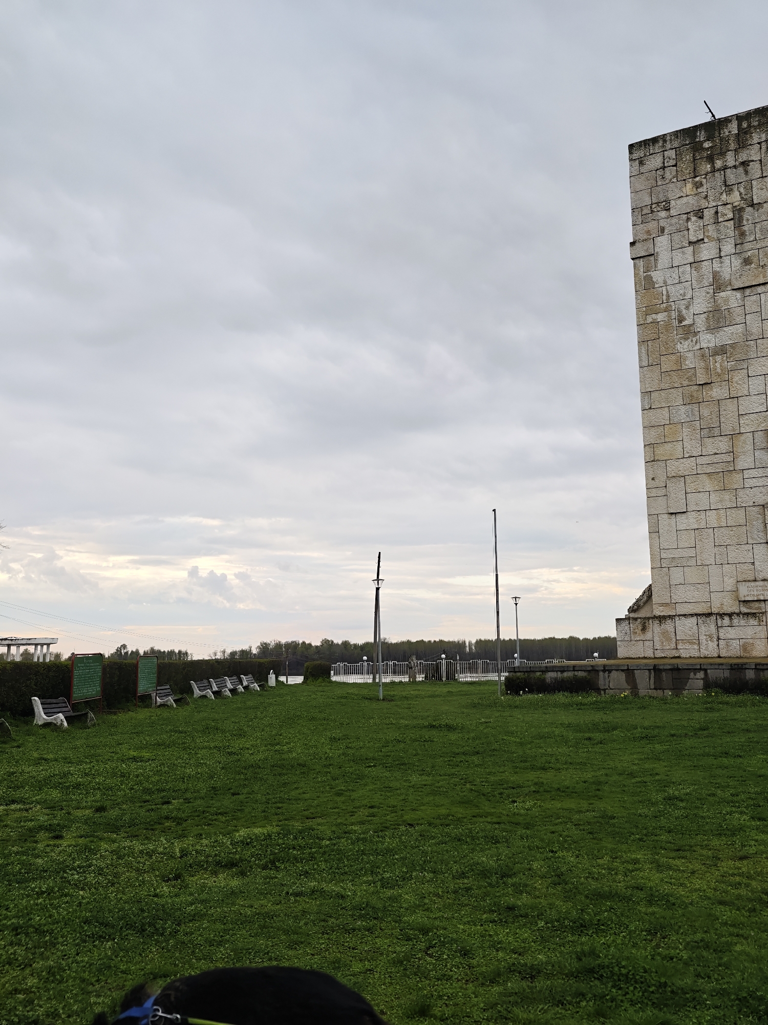 The image shows a grassy area next to a large stone building, possibly a monument or memorial, under a cloudy sky. In the distance, there is a body of water and a bridge-like structure. There are several benches and informational signs along the edge of the grass. The bottom of the frame shows a dark object with a blue collar, possibly a dog. The location is likely in the city of Kozloduy, Bulgaria.