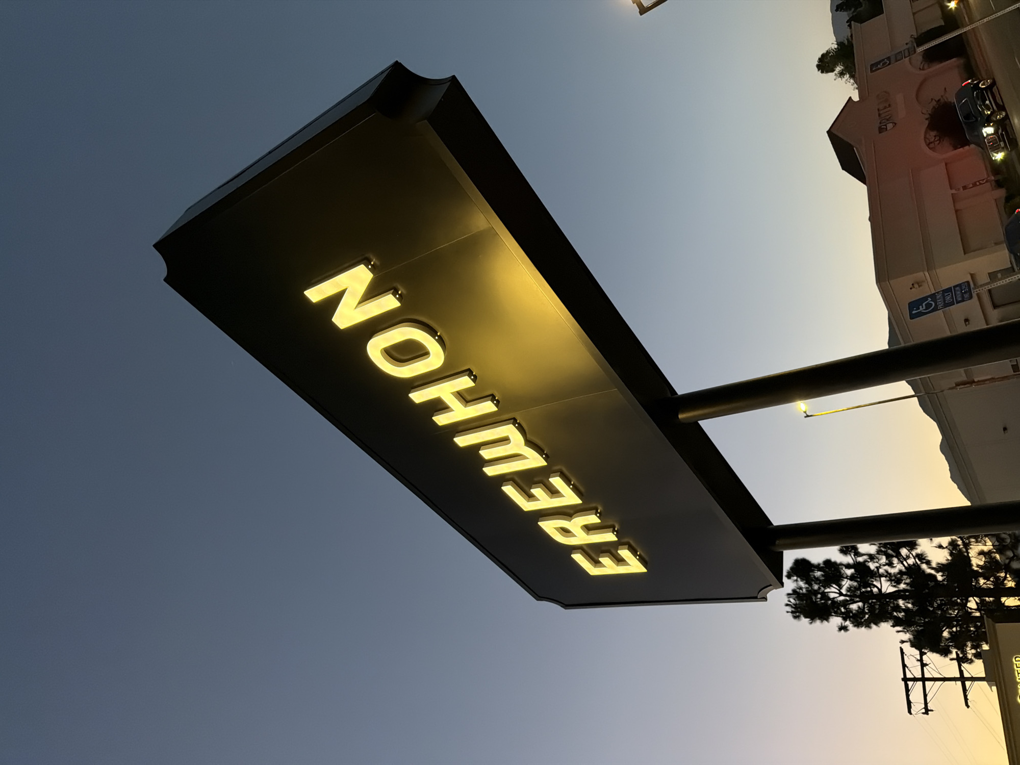 The image depicts a sign with the illuminated word "EREWHON" against a dusky sky. The sign is large and dark with bright yellow lettering. Two thick black posts support the sign, which is angled upwards. In the background, a silhouetted tree stands to the left, and a building with the word "RITE AID" visible on its facade is to the right. The overall environment suggests an urban or suburban setting at dusk. The location is identified as Glendale, United States of America.