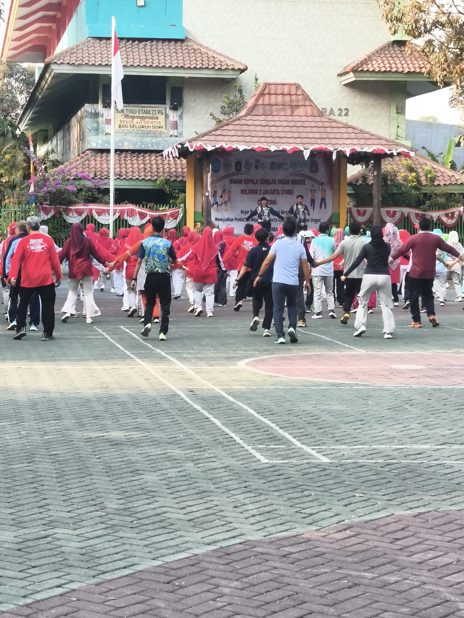 The image depicts an outdoor gathering in front of a school building in North Jakarta, Indonesia, likely for a school event. In the foreground, a group of people, predominantly students and teachers, are engaged in a synchronized exercise routine on a paved court marked with white lines. Many participants are wearing red or brightly colored athletic wear and are holding hands, suggesting a communal activity like aerobics or a warm-up.
Behind the participants is a small stage or platform adorned with red and white decorative trim, and a banner that appears to have Indonesian text, likely related to the event's title or purpose. Two individuals are visible on the stage, possibly instructors or performers. The school building in the background has a light blue facade and a distinctive tiled roof. A flag pole with an Indonesian flag is prominently positioned to the left of the stage. The environment is sunny, with trees visible in the upper right corner. The overall scene conveys a sense of community and school spirit, with a focus on physical activity.