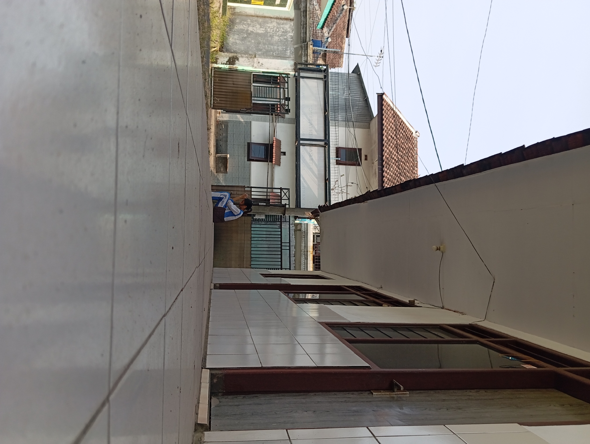 An exterior, low-angle shot shows a tiled walkway leading towards a building with a covered walkway. The covered walkway has a white ceiling and is supported by columns. The building's facade is tiled and has a series of windows with dark brown frames. A young person wearing a blue and white uniform is sitting on a dark object, facing away from the camera, with their head bowed. In the background, there are other buildings with tile roofs, some with corrugated metal extensions, and power lines. The sky is overcast. The location is confirmed as Pandaan, Indonesia.