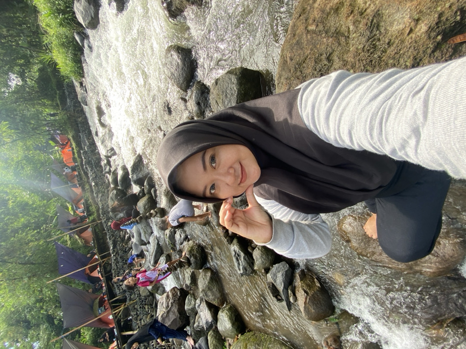 A woman wearing a hijab poses for a selfie while standing in a rocky stream. Clear water flows around her feet and through the rocks. Behind her, a row of tents suggests a campground beside the stream. Other people are visible further down the stream, wading or playing in the water. The scene is bright and sunny, with green foliage visible along the bank.