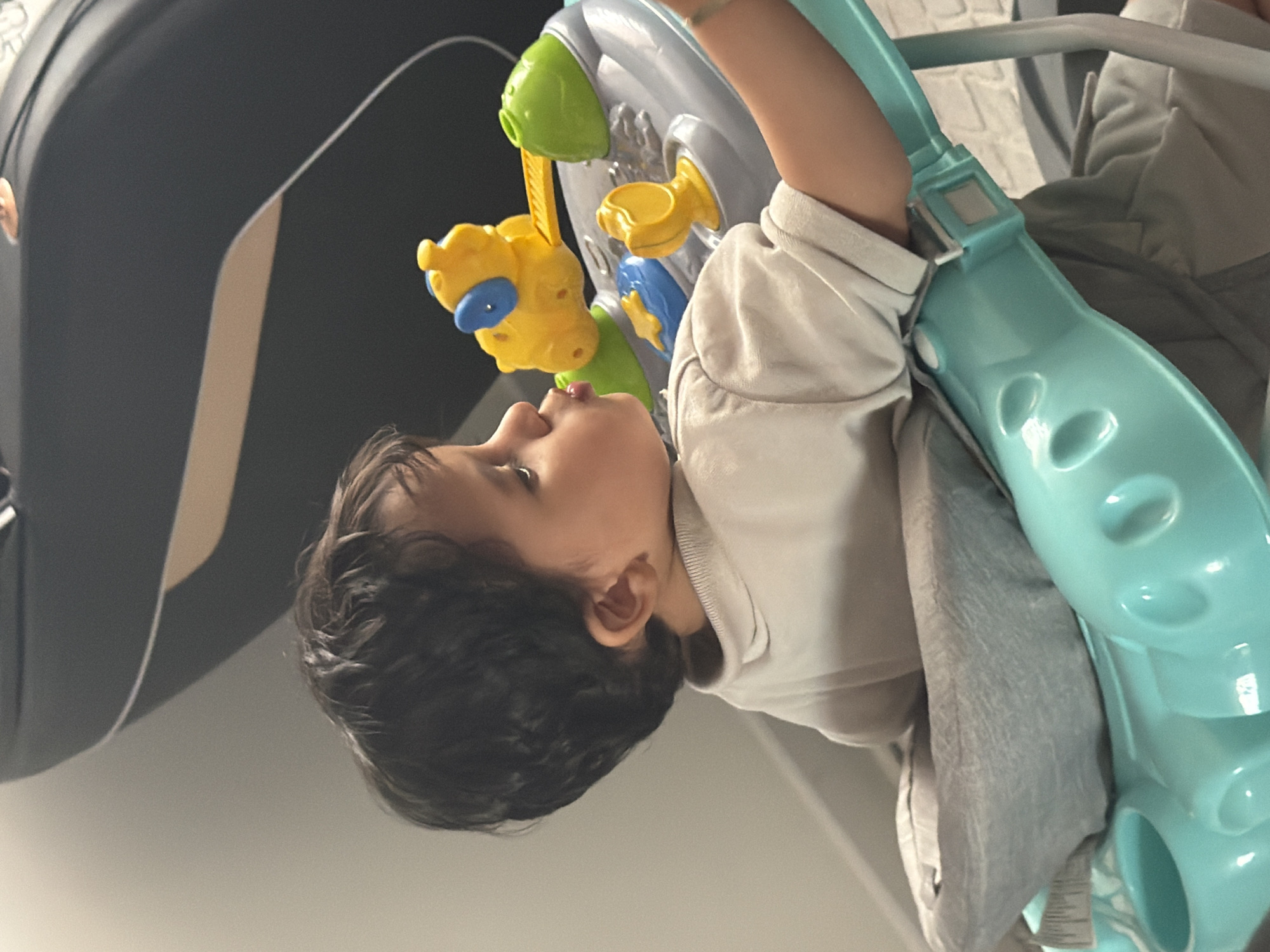 The image displays a young child, appearing to be a toddler, seated in a multi-colored activity center within an indoor setting. The child is positioned in profile, facing the right side of the frame, with dark, curly hair and a light to medium skin tone. They are wearing a light beige or off-white short-sleeved top and light-colored bottoms. Their lips are distinctly pursed, indicating focus or an action like blowing. A faint, light-colored band or bracelet is visible on the child's right wrist.
The activity center features a grey-colored main tray adorned with several brightly colored plastic toys. Notable toys include a yellow and blue animal-shaped interactive component with a yellow lever, a green domed toy, a yellow U-shaped handle, and a blue star shape. The base structure of the activity center is a prominent teal or turquoise color, with a grey fabric seat supporting the child.
The background consists of a plain light grey wall to the left of the child. To the upper right, a large, dark grey or black upholstered furniture piece with a distinct light tan or cream-colored curved accent line is partially visible, suggesting a modern design. The lighting is even and consistent with an interior space. No specific geographical location, such as the city of Sinitovo, Bulgaria, can be determined from the visual information provided in the image.