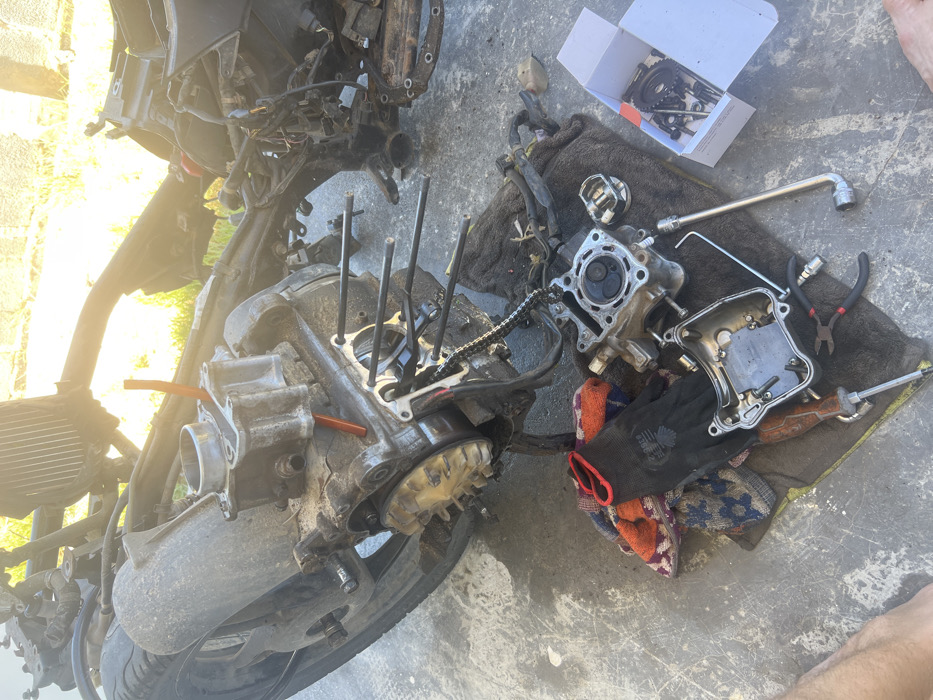 This image captures a motorcycle engine in a state of advanced disassembly on a concrete surface in Cam Đức, Vietnam. The main engine block, heavily soiled, is resting on the ground with its cylinder head removed. Four long cylinder studs protrude from the crankcase, and a timing chain is visible. A detached cylinder block, containing a liner, rests on the engine casing, with an orange plastic tube inserted into it. To the left, a flywheel or stator assembly is partially visible.
Adjacent to the engine, various components and tools are scattered on rags. A cylinder head, displaying its valves and combustion chamber, is positioned on a dark rag alongside a piston and a separate valve cover. Among the tools present are a screwdriver with a wooden handle, a pair of pliers, an Allen wrench, and a socket wrench extension. A multicolored patterned rag is also visible. A white cardboard box, open, contains an assortment of smaller metallic engine parts, including gears and bolts.
The background shows the stripped frame of a scooter or motorcycle, including what appears to be a radiator, with dense green foliage illuminated by bright sunlight beyond. The scene takes place outdoors on a textured gray concrete slab. The lower left corner of the image reveals a bare human foot, indicating the presence of an individual engaged in the repair or maintenance activity.