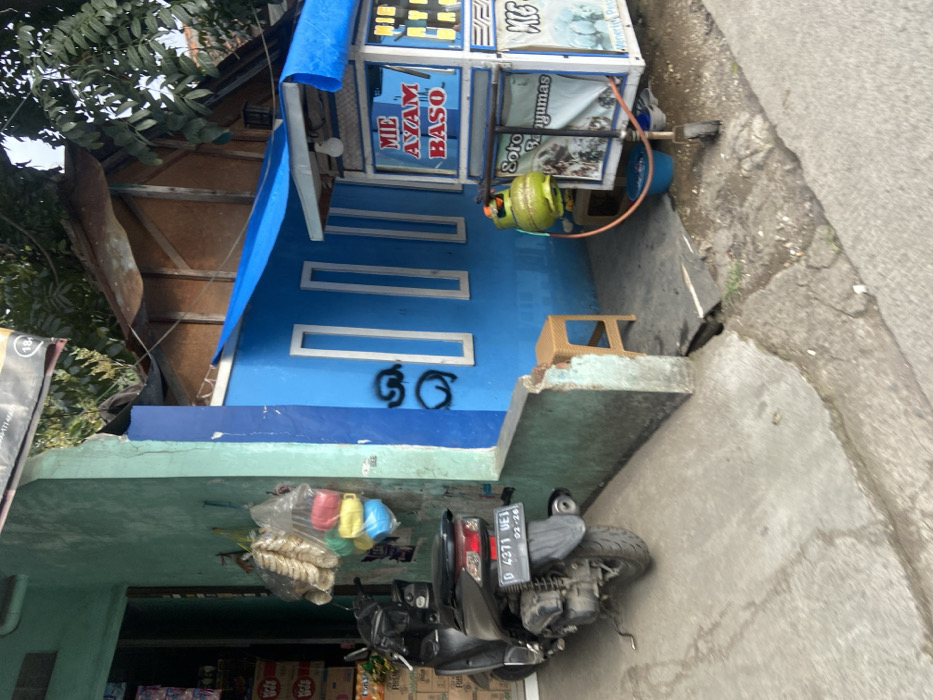 A black scooter with the license plate "D 4271 WEJ 02-26" is parked in front of a green and blue building in Margaasih, Indonesia, at coordinates -6.961, 107.546. The building is painted blue with a green trim. There are three windows on the blue wall and graffiti is visible on the wall below the windows. A yellow propane tank is next to a food cart, which is located on the sidewalk to the right of the scooter. The food cart has a blue tarp roof and says “Mie Ayam Baso" in red letters. There is a wooden stool in front of the blue wall. There is also a small blue plastic basin in front of the food cart. The sidewalk is made of concrete and is cracked in places. The scene is lit by daylight.