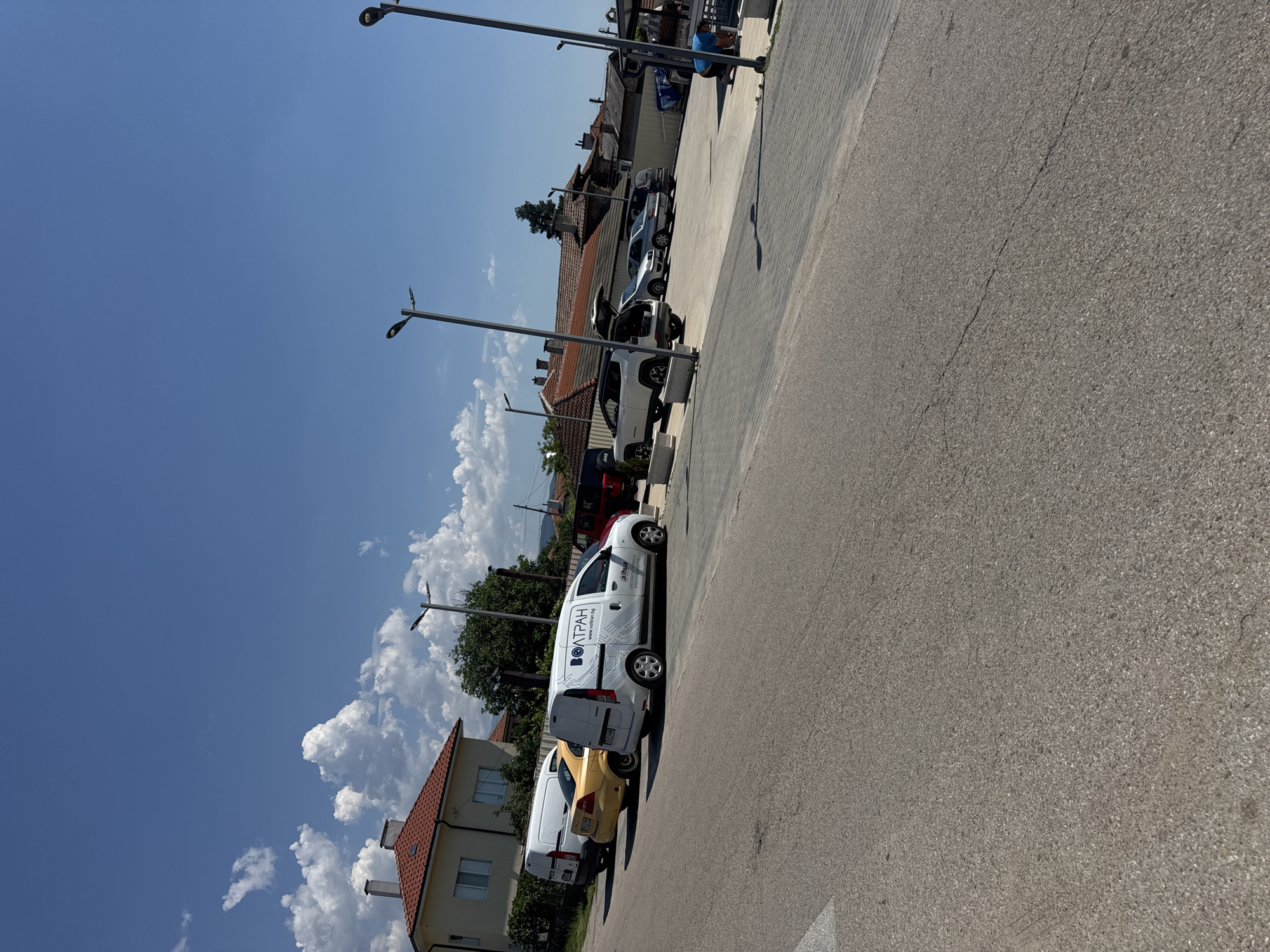 The image shows a street scene in Vedrare, Bulgaria, on a sunny day. In the foreground, a paved road curves to the right. In the middle ground, several vehicles are parked on a paved lot. These include a white van with the logo "ВОЛТРАН" and "www.voltran.bg", a yellow car, a white van, a white Jeep with its trunk open, and other cars. The background features houses with red tiled roofs, trees, and streetlights. The sky is blue with scattered white clouds. A person in a blue shirt is visible in the far right of the image, appearing to be tending to something on the ground.