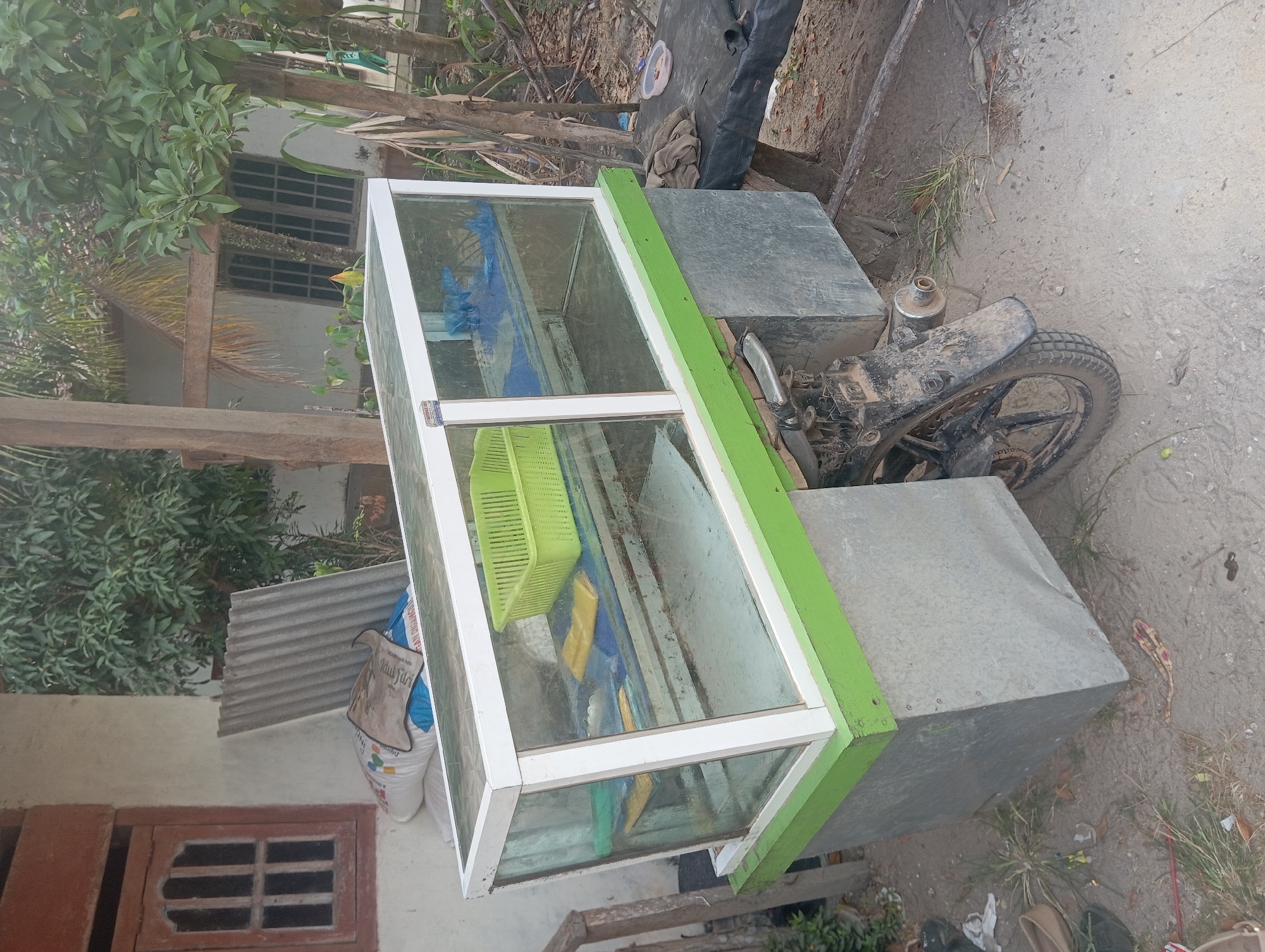 The image shows a modified motorcycle with a glass display case mounted on its rear. The motorcycle appears to be stationary and is parked on a dirt or gravel surface. The display case is rectangular with a white frame and appears to be made of glass. It has a green base and is filled with a few items, including a green plastic basket and some yellow and blue objects. On the left side of the motorcycle, there are large galvanized metal boxes attached. In the background, there is a building with a window and a corrugated metal roof, along with trees and vegetation. The scene suggests an outdoor market or street vendor setup in a rural or semi-urban environment. The specific location is Dente Teladas, Indonesia, as indicated by the context.