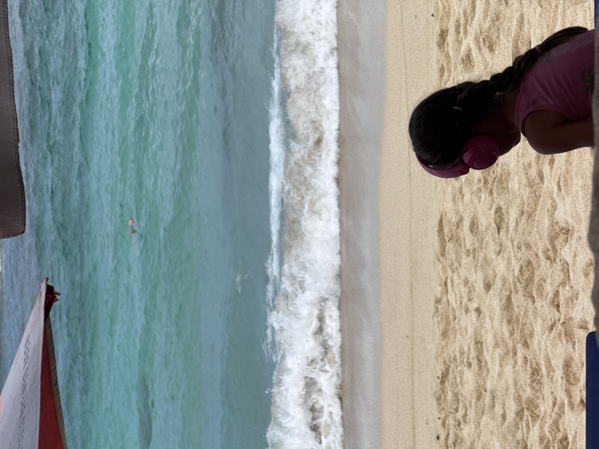 A sideways shot shows a beach scene with a child wearing pink headphones on the sand. The shoreline is visible with crashing waves, and turquoise water stretches out to the horizon. A person is swimming in the ocean. A red and white object is partially visible in the lower left corner. The location appears to be a beach in Kuta Selatan, Indonesia.