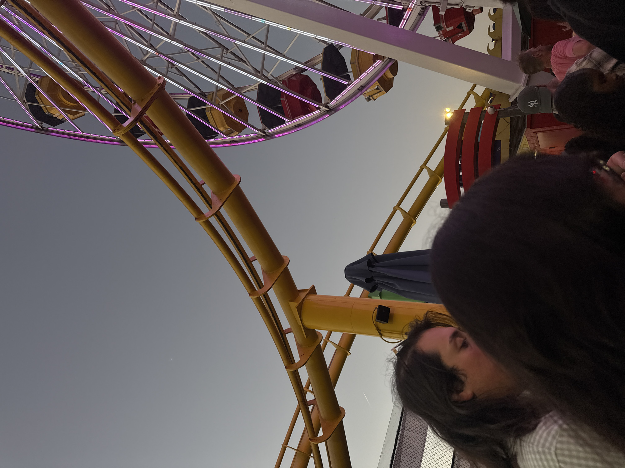 The image shows a view of a Ferris wheel and a roller coaster at dusk in Santa Monica, California. The sky is a muted gray, with a hint of twilight. The Ferris wheel, adorned with colorful lights, is in the right portion of the frame, its wheel spokes and gondolas visible. The roller coaster track, painted a bright yellow, curves across the upper left of the image. In the foreground, several people are visible from the shoulders up, their faces partially obscured, suggesting they are part of a crowd or are seated. One person in the middle ground is wearing a gray baseball cap with a white logo. The scene appears to be at an amusement park or pier, given the presence of these rides and the general atmosphere.