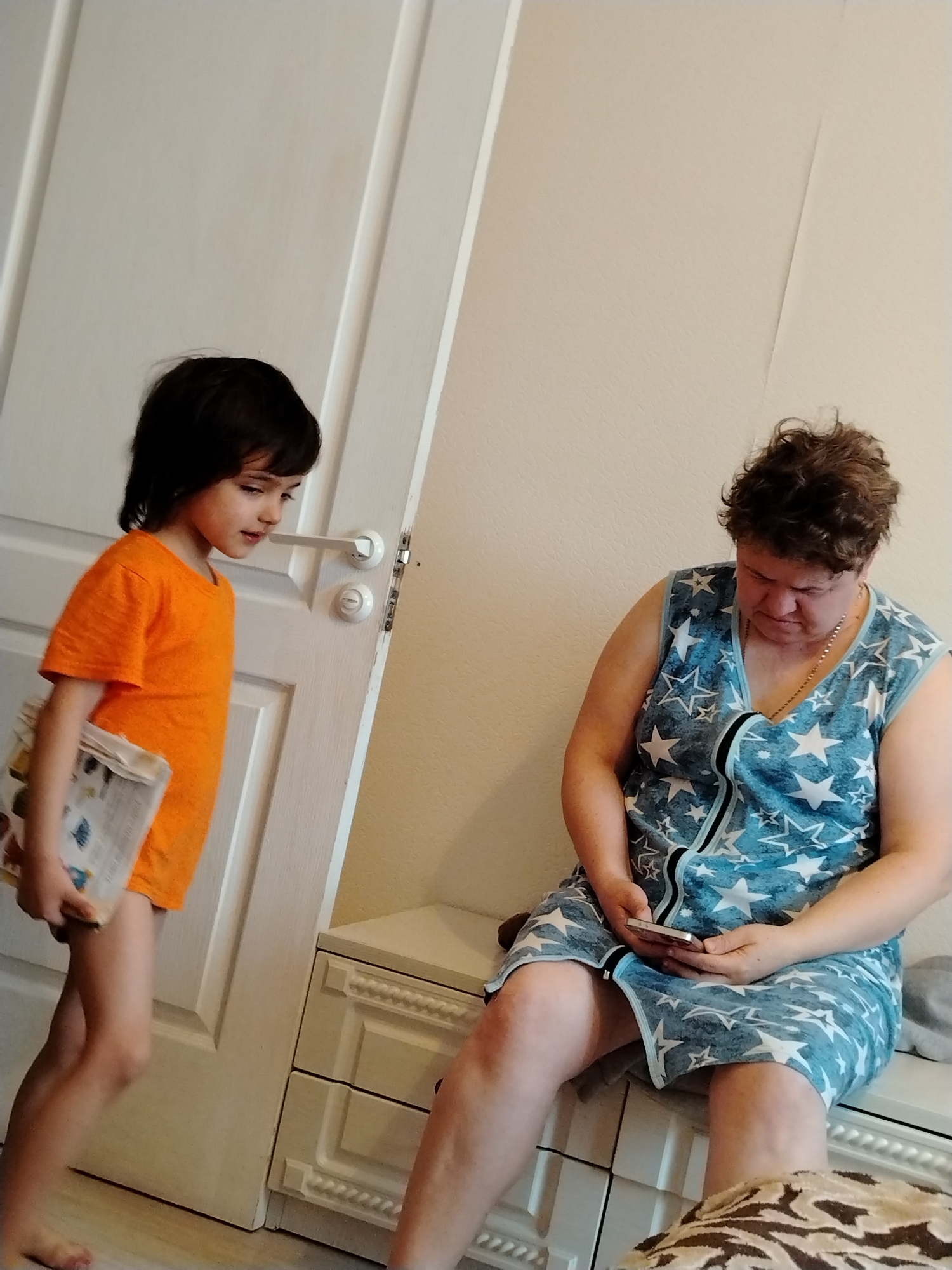 A young child stands in front of a closed white door, holding a book and wearing an orange t-shirt. An older woman sits on a white dresser, looking at a cell phone. She is wearing a light blue robe with white star patterns. The setting appears to be inside a residence. The location is not explicitly stated as Noviy, Russia.