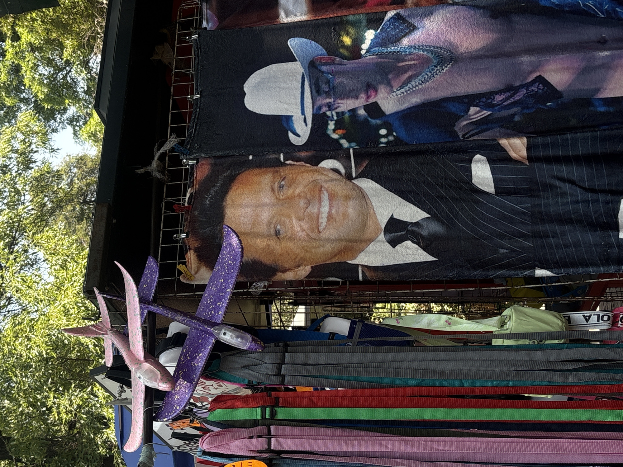 This image captures a vibrant outdoor market stall, likely in Mexico City, displaying various merchandise under bright daylight. Prominently featured are two large tapestries or blankets hanging from a metal grid display rack.
The lower tapestry depicts a smiling Luis Miguel, wearing a pinstripe suit and a black bow tie. Above him, another tapestry features a person with a white cowboy hat, a dark shirt, and a notable beaded necklace or chain, presented in a stylized manner.
Below these tapestries, a diverse array of colorful items is displayed. Two foam toy airplanes, one pink and one purple, hang suspended; the purple plane has a small light near its nose. Further down, numerous straps or belts in various colors, including red, green, blue, pink, grey, and black, are stacked or draped, likely offered for sale. A light green bag or pouch is partially visible behind these straps. The tapestries are held in place by binder clips.
The background shows lush green trees against a bright sky, indicating a sunny, clear day in an outdoor setting, possibly a park or an open-air market. Part of the word "OLA" is visible on a white strap in the lower right section of the image. The scene suggests a lively commercial activity with items targeted at a popular audience.