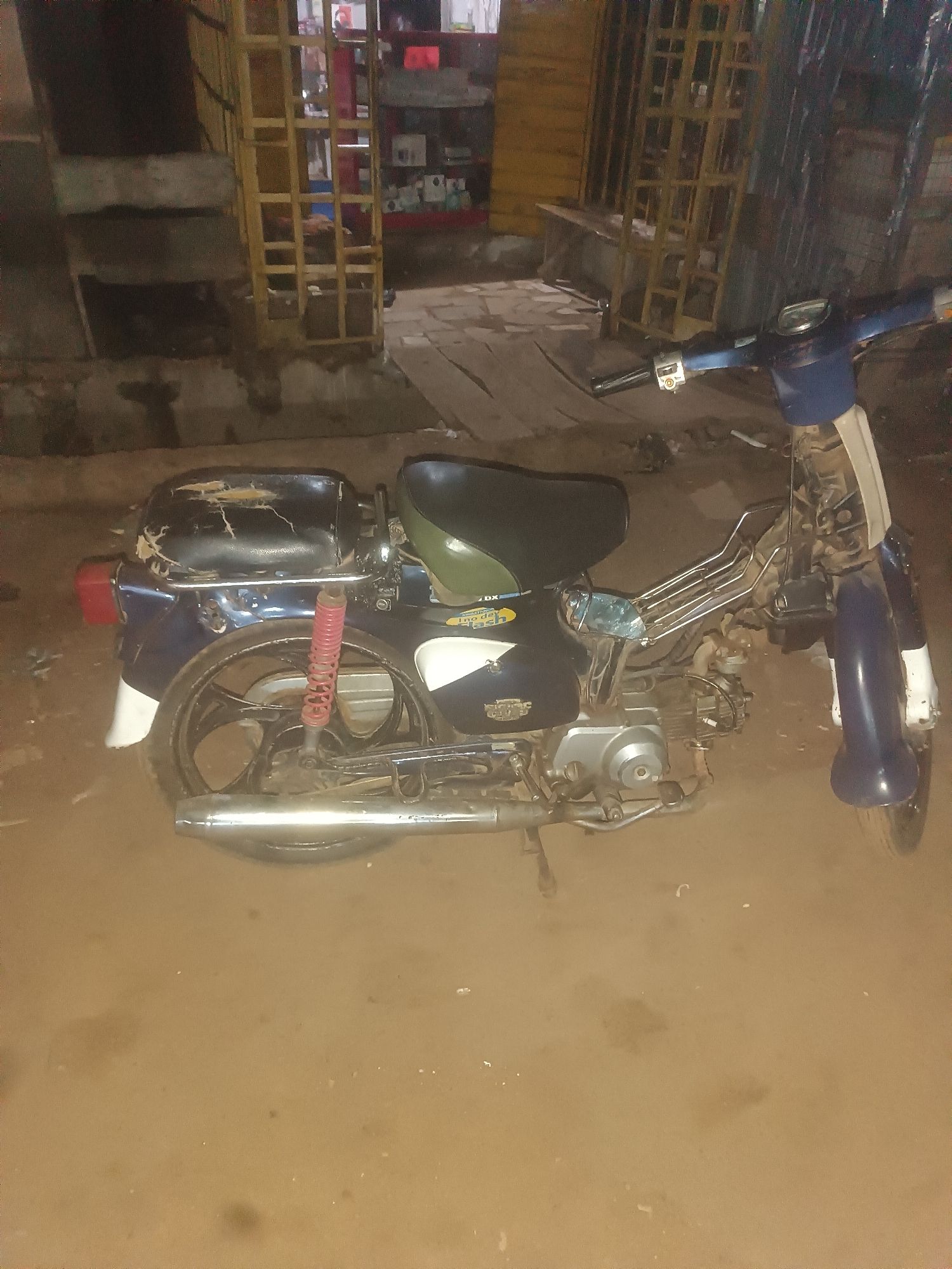 An old, blue and white moped is parked on a dirt ground in front of a store with yellow metal shelves in Wukari, Nigeria. The moped is weathered, with a torn black seat and a red rear shock absorber. The store behind it has shelves stocked with various items, though they are not clearly identifiable due to the low lighting and image quality. The ground is uneven and dusty, suggesting an outdoor, possibly informal, setting.