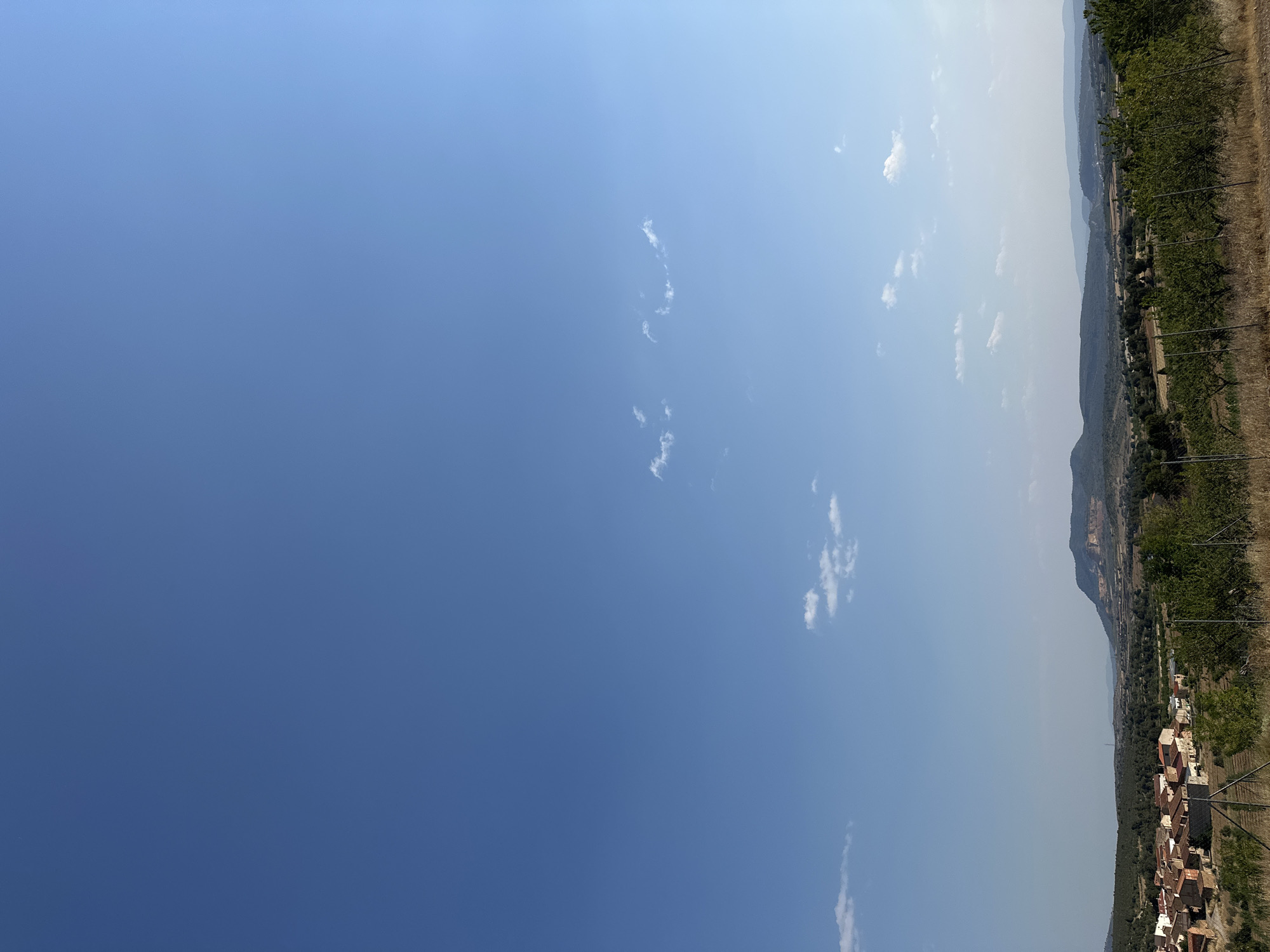 The image depicts a panoramic view of the town of Alpuente, Spain, under a clear blue sky with scattered white clouds. In the foreground, a fence separates the viewer from dry, grassy terrain and the edge of a vineyard or orchard with green trees. Beyond this, the town of Alpuente is visible, characterized by a cluster of buildings with terracotta roofs and stone walls. The town is nestled amidst a landscape of rolling hills covered in green trees and vegetation. In the background, a prominent mountain range rises, with visible geological strata and a distinct plateau shape. The overall scene is one of a rural, sun-drenched environment.