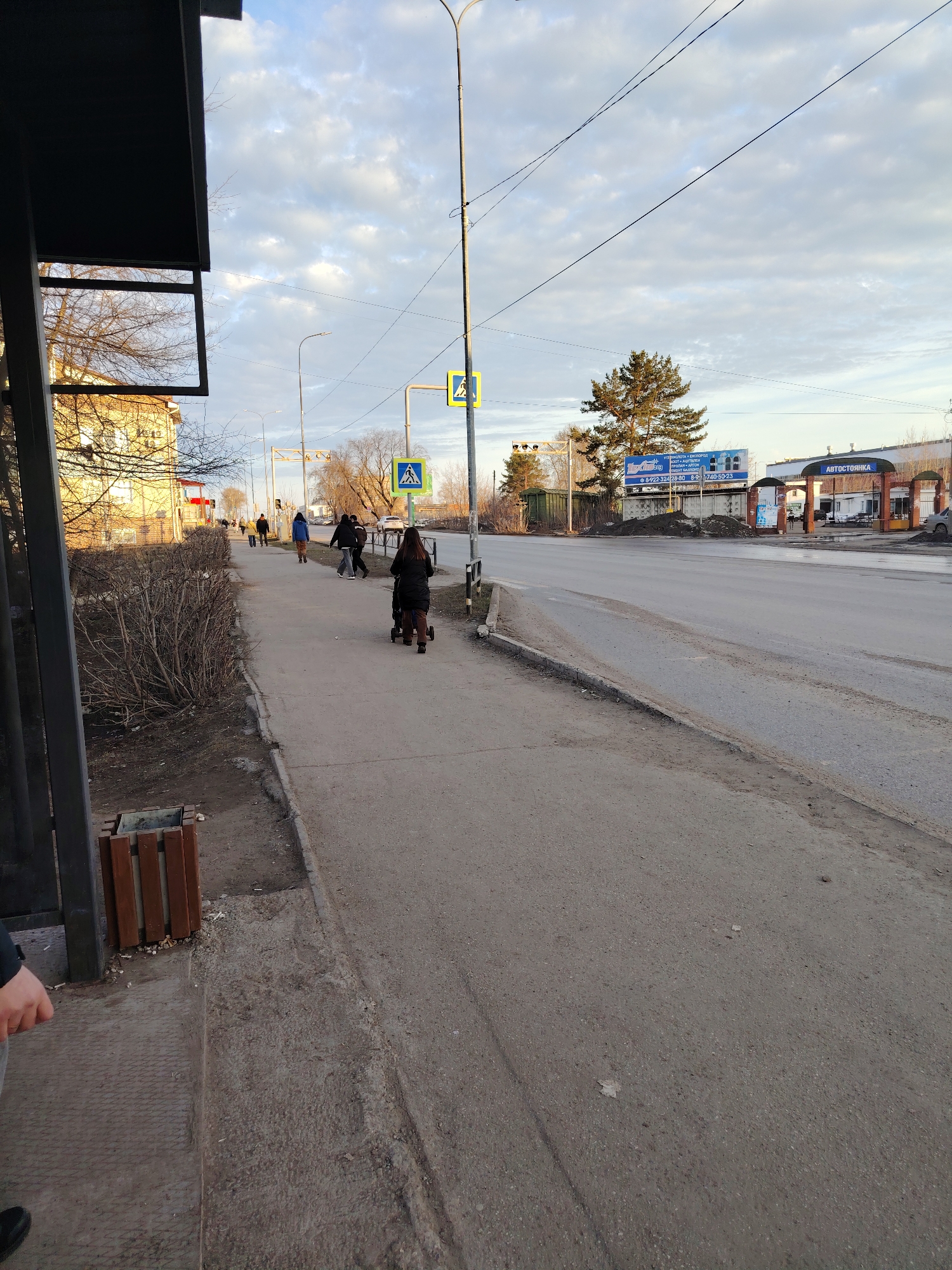 The image depicts a street scene in Chaikovsky, Russia, showcasing a pedestrian sidewalk adjacent to a roadway. Several pedestrians are walking along the sidewalk, including a woman with a stroller. Road signs are visible on the side of the road, marking a pedestrian crossing. Across the street is an "автостоянка", which translates to "parking lot" in English. The overall setting appears to be an urban area with a mix of commercial and residential buildings in the background. The weather seems overcast, with a cloudy sky above. A bus stop with a trash receptacle is on the left.