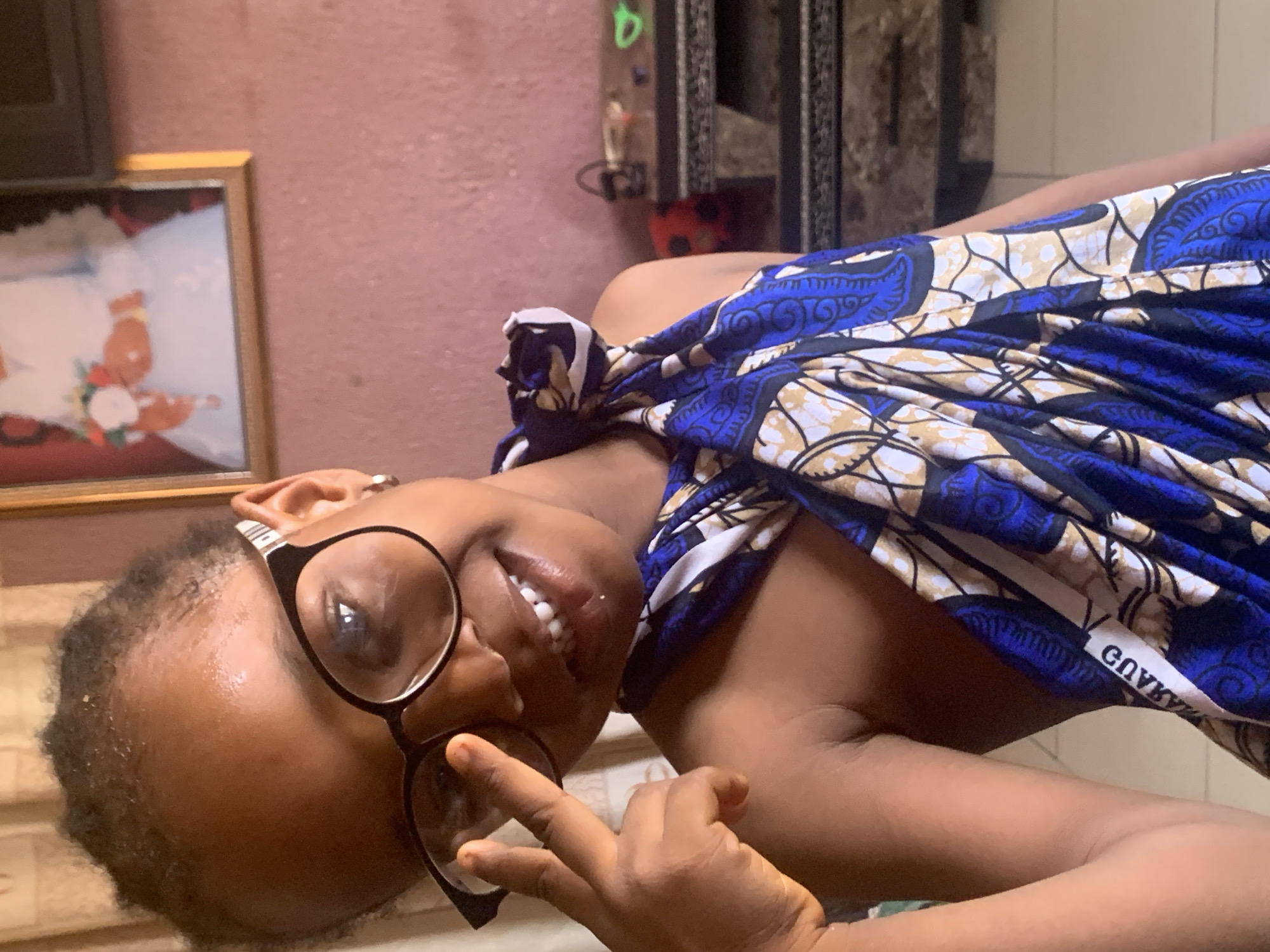 A woman with dark skin and short hair is seen lying on her back, wearing large, round black glasses. She has a blue and beige patterned cloth draped over her upper body. Her right hand is raised, with her index and middle fingers extended in a peace sign, partially obscuring her glasses. She is smiling, revealing her teeth.
In the background, a framed picture hangs on a pink wall. A partially visible dresser or cabinet stands behind her.
It is not possible to confirm if the image was taken in the city of Sabon Tasha, Nigeria.