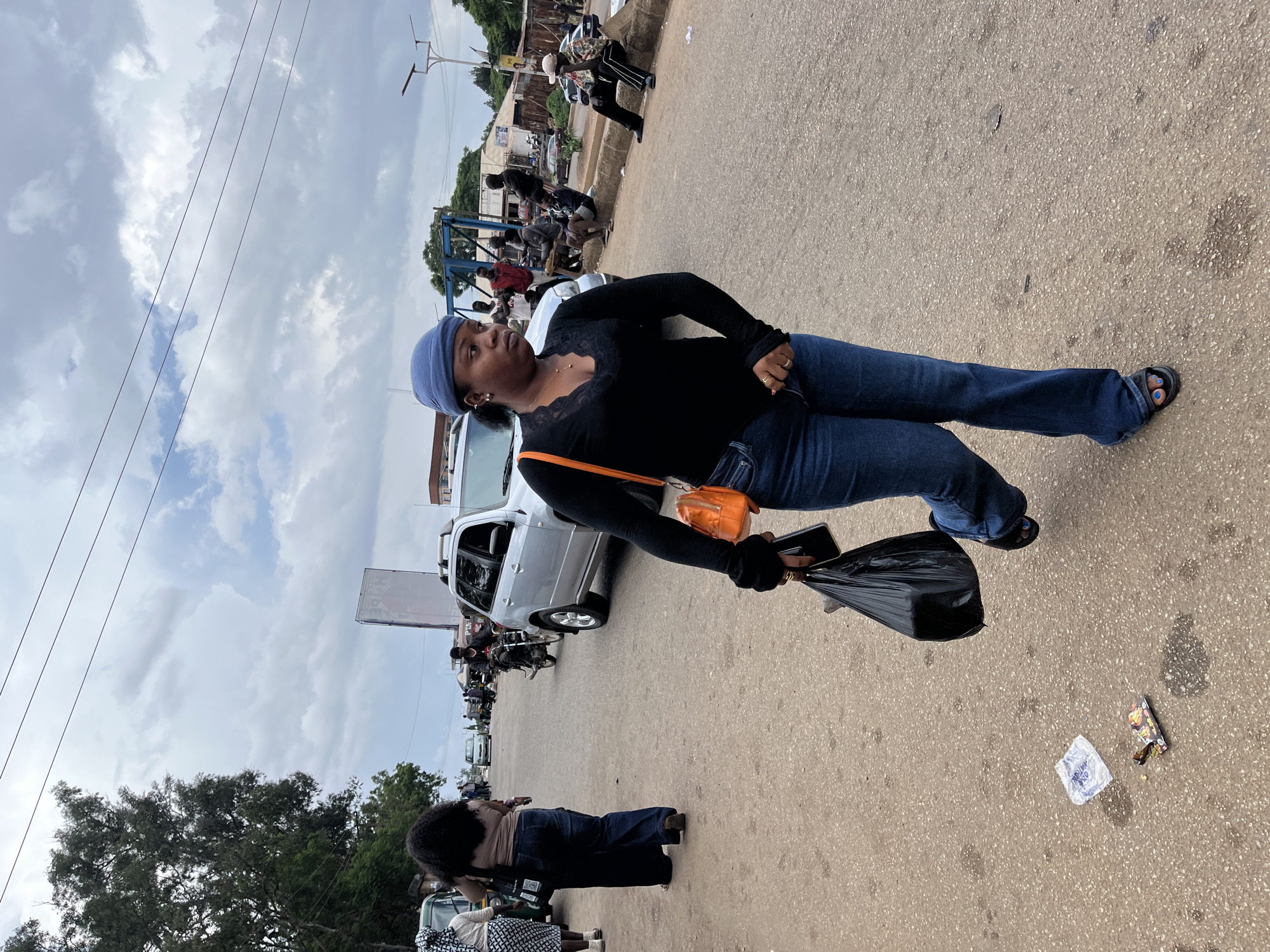 The image depicts a street scene in Jos, Nigeria, under a cloudy sky. The primary subject is a Black woman standing on a paved road, wearing a blue headscarf, a black long-sleeved shirt with lace detailing at the neckline, and dark blue jeans. She is carrying a black plastic bag in her right hand and a smartphone in her left. An orange purse is slung across her body. She appears to be looking off to the side, her expression neutral.
The setting is a busy street with vehicles, including a silver SUV parked nearby, and motorcycles. Several other individuals are visible in the background, some standing and others appearing to be seated or working. The road is a mix of asphalt and concrete, with some debris and litter scattered on the ground, including a crumpled piece of white paper and what looks like a small food wrapper.
Further down the street, a mix of trees and buildings suggests an urban or semi-urban environment. The overall impression is of a typical day on a Nigerian street, likely a market or commercial area, given the presence of many people and vehicles. The weather appears overcast, possibly hinting at a time of day when the sun is not directly overhead, though the specific time is not discernible. No visible text is clearly legible in the image.
