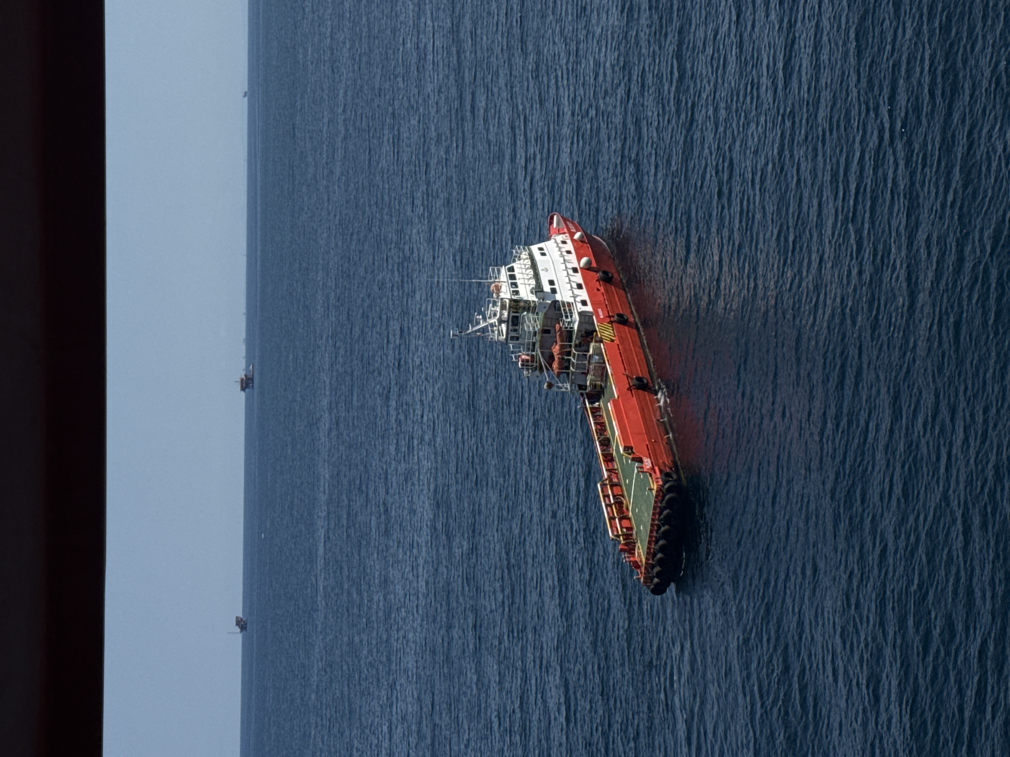 A red and white offshore supply vessel is sailing on a calm, deep blue ocean. The vessel has a large red deck at the stern and a white superstructure at the bow. In the distance, on the horizon, are several oil rigs and smaller vessels, suggesting an industrial offshore area. The sky is a clear, light blue. The image is taken from a high vantage point, possibly another vessel or a structure. The location is identified as Unknown, Italy.