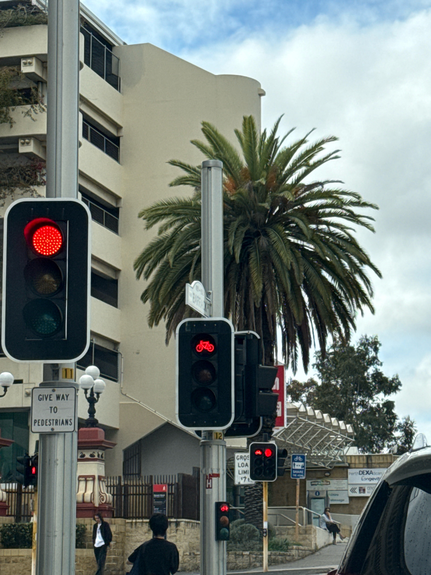 The image shows a street scene in an urban environment. A traffic light with a red light illuminated is visible on the left side of the frame, mounted on a metal pole. To the right of this, another traffic signal displays a red bicycle symbol, indicating a bike lane signal. Further to the right, a street sign with the text "BARRA ST" is affixed to a pole. Behind the traffic signals, a multi-story building with a light-colored facade and balconies is visible. A large palm tree with green fronds stands prominently in the middle ground. In the foreground, a portion of a silver vehicle is visible on the right edge of the frame. Two individuals are present on the sidewalk; one, wearing dark clothing, walks away from the camera, while another, in dark clothing, faces away from the camera. A "GIVE WAY TO PEDESTRIANS" sign is mounted on the pole supporting the main traffic light. The sky is cloudy.