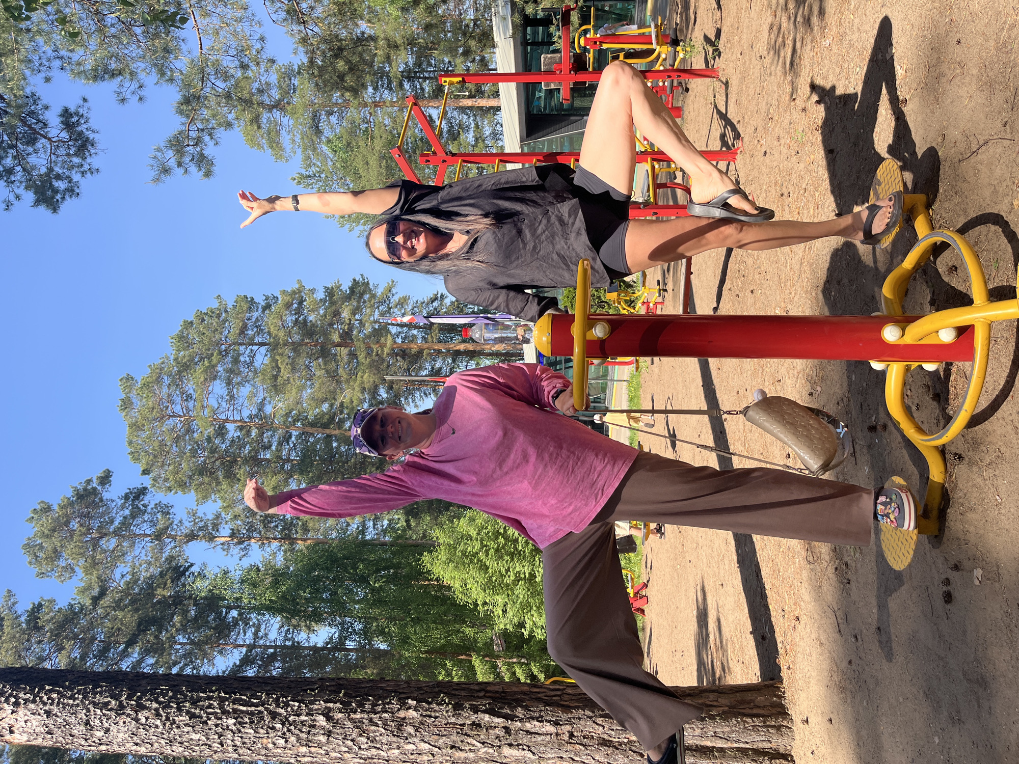 Two women are posing on outdoor exercise equipment in a park. The woman on top is wearing a black shirt, black shorts, sunglasses, and sandals. She has one arm raised in the air. The woman on the bottom is wearing a pink shirt, brown pants, a hat, and sandals. She also has one arm raised in the air. The exercise equipment is red and yellow. The park has trees and other exercise equipment in the background. The location is Chaykovsky, Russia.