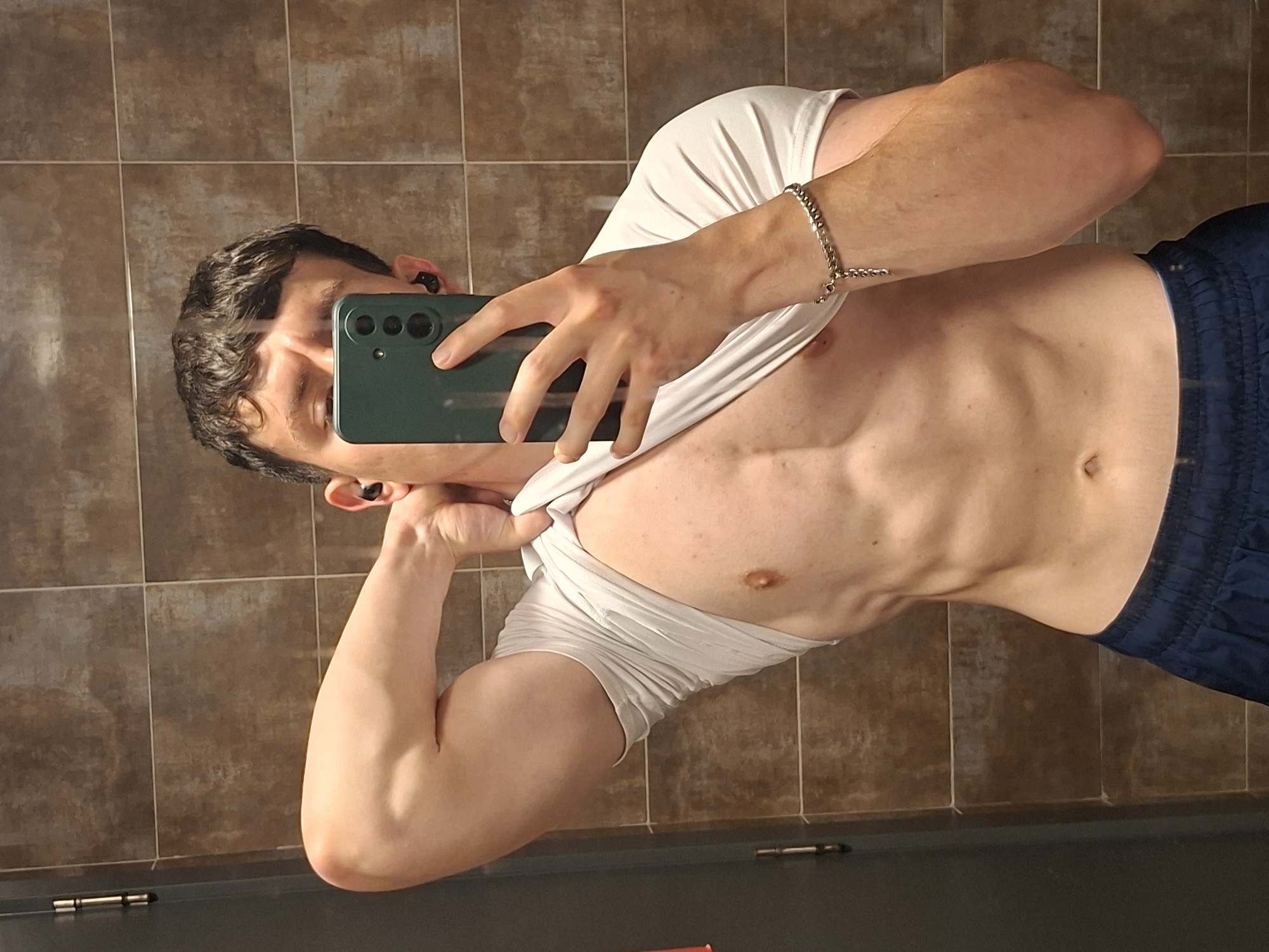 A person is taking a selfie in what appears to be a bathroom with brown tile walls. The individual, a man, is holding up a green smartphone in his right hand to capture the image. He is wearing a white t-shirt which is partially lifted to reveal his torso. Blue shorts are also visible. A silver bracelet is on his right wrist. The man has short dark hair, and is wearing black earbuds. The location cannot be determined to be Plovdiv, Bulgaria from this image alone.