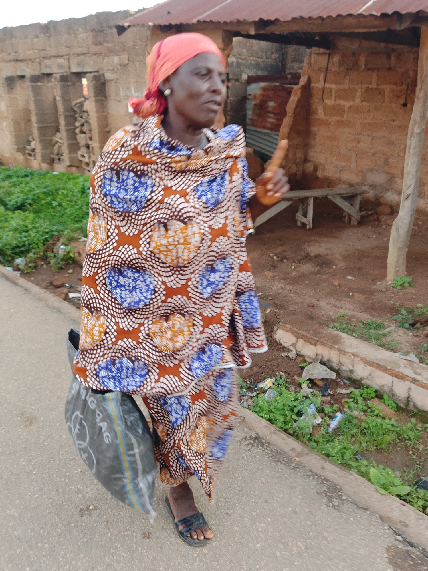 A full-shot, eye-level outdoor photograph captures an elderly woman with dark skin, wearing an orange headscarf and a colorful patterned wrap dress, walking along a paved street in Jos, Nigeria. The woman is facing to her right and looking slightly upward. In her right hand, she holds a small, light brown object. A dark gray plastic bag hangs from her left arm. She wears black sandals on her feet.
The background consists of a weathered, brick building with an corrugated metal roof and a wooden bench in front of it. The ground around the building is unpaved and covered with dirt and vegetation. The street is lined with a concrete curb, and to the right of the curb, there is a drainage ditch filled with green plants and scattered trash, including plastic bottles. The overall impression is one of an urban environment with signs of both habitation and neglect.