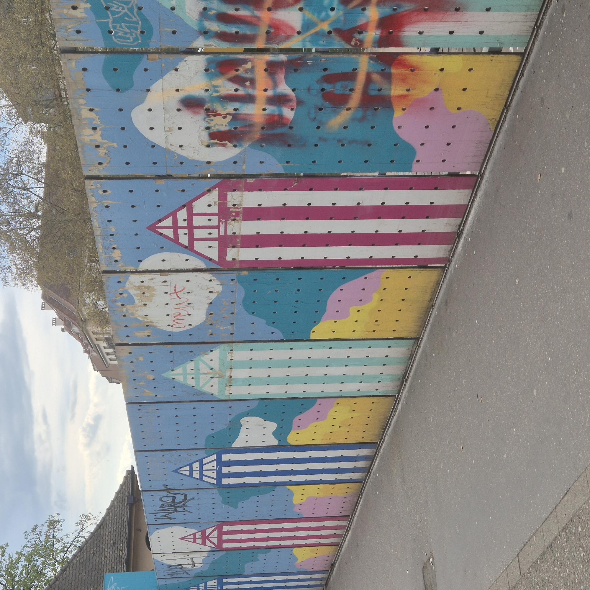 The image shows a colorful, abstract mural on a wall made of wooden planks in Strasbourg, France. The mural features repeating motifs of striped beach huts with triangular roofs, painted in various color combinations: pink and white, light blue and white, and dark blue and white. The background is predominantly blue with cloud-like shapes in white, yellow, and pink. Some planks have graffiti tags in black. The planks are weathered and have rows of evenly spaced holes. The wall runs parallel to a gray concrete pavement. Parts of buildings with brick and tiled roofs are visible in the background, as well as trees with budding leaves and a cloudy sky.