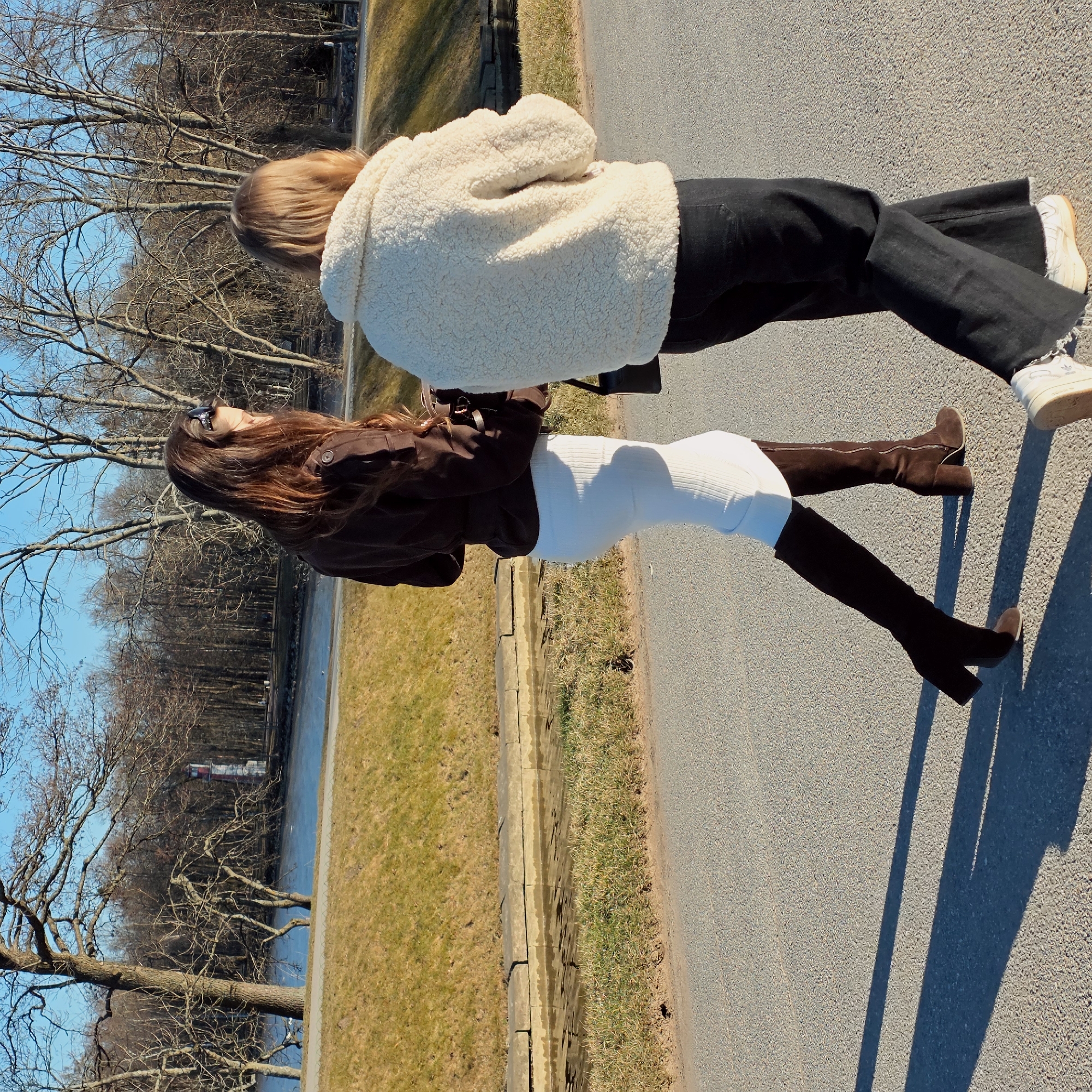 Two women stand on an asphalt path in Peterhof, Russia. One, in a brown jacket, white dress, and brown knee-high boots, looks toward the other, who wears a white fuzzy jacket, black pants, and white sneakers. The scene includes a park-like setting with bare trees, grass, and a body of water. The bright sunlight casts shadows, indicating a clear day.