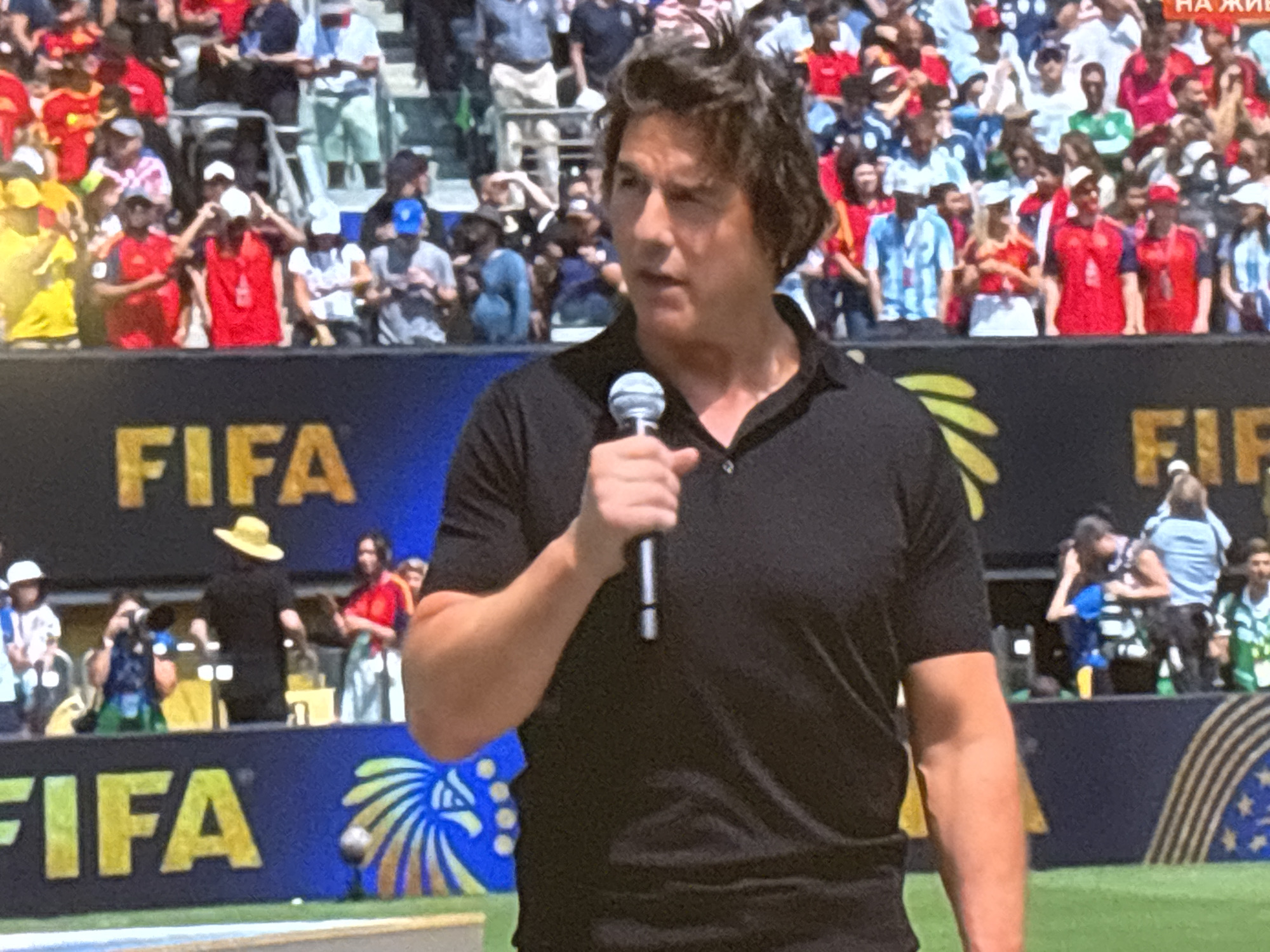 The image shows a man, identified as Tom Cruise, holding a microphone and speaking at an outdoor event. He is wearing a black polo shirt. Behind him, a crowd is visible in tiered seating, many wearing red and yellow attire, suggesting a sporting event. The word "FIFA" is emblazoned on a blue banner in the foreground. The background includes more spectators and a large screen or backdrop. The scene is brightly lit, indicating daytime. While the prompt requests a location in Burgas, Bulgaria, there are no visual cues in the image to confirm this specific location.