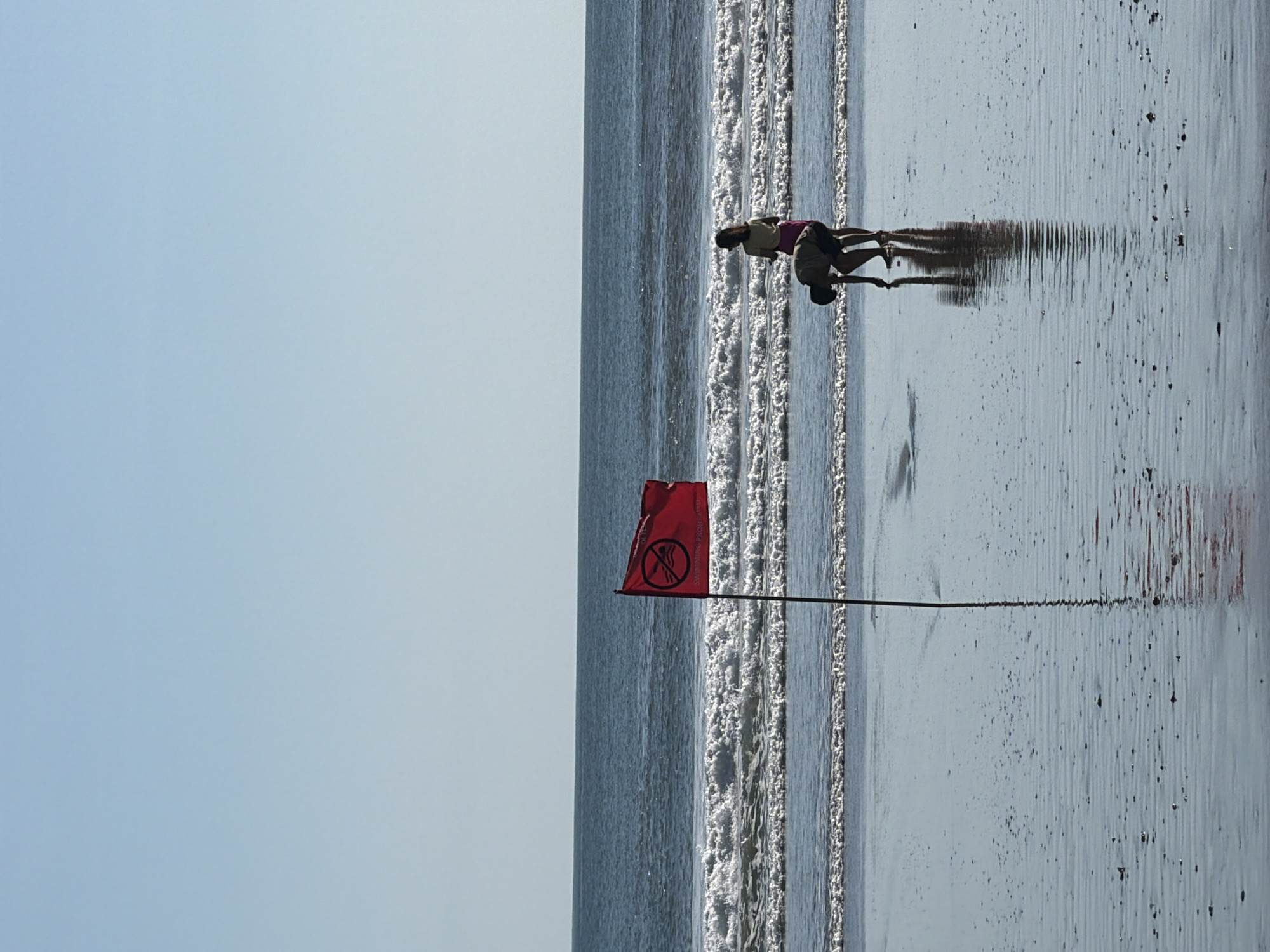 Two individuals are on a beach in Kuta, Indonesia. The person in the foreground is wearing a pink top and shorts, while the person behind them is wearing a dark top and shorts. They appear to be interacting in the shallow water near the shore, possibly wrestling or playing.
A red flag with a black symbol is prominently displayed on a pole. The symbol on the flag appears to be a circle with a diagonal line through it, and inside the circle is a depiction of a person swimming. This is a common symbol indicating that swimming is prohibited. The flag is positioned near the waterline.
The scene takes place on a beach with wet sand reflecting the sky and waves. The ocean is calm with small waves breaking near the shore. The sky is clear and blue, suggesting a sunny day, likely in the afternoon given the angle of the shadows. The wet sand shows ripples from receding water and scattered dark specks, possibly shells or debris. The overall atmosphere is relaxed and summery.
