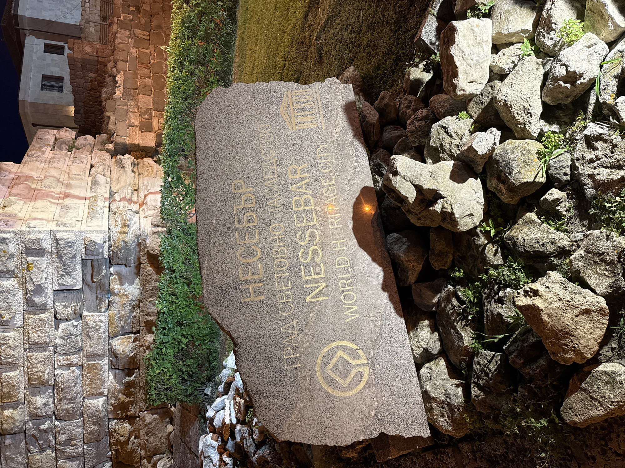 The image shows a stone monument in Nesebar, Bulgaria, that reads "NESSEBAR" in both Cyrillic and Latin alphabets, and "WORLD HERITAGE CITY". The monument is situated in front of a stone wall and a manicured green hedge. The setting appears to be a historical site, possibly an ancient ruin, given the stonework of the background structures. The lighting suggests it is nighttime, with artificial lights illuminating the scene. There are no people or animals visible in the image. The monument is made of rough-hewn stone and rests on a bed of smaller rocks and grass.