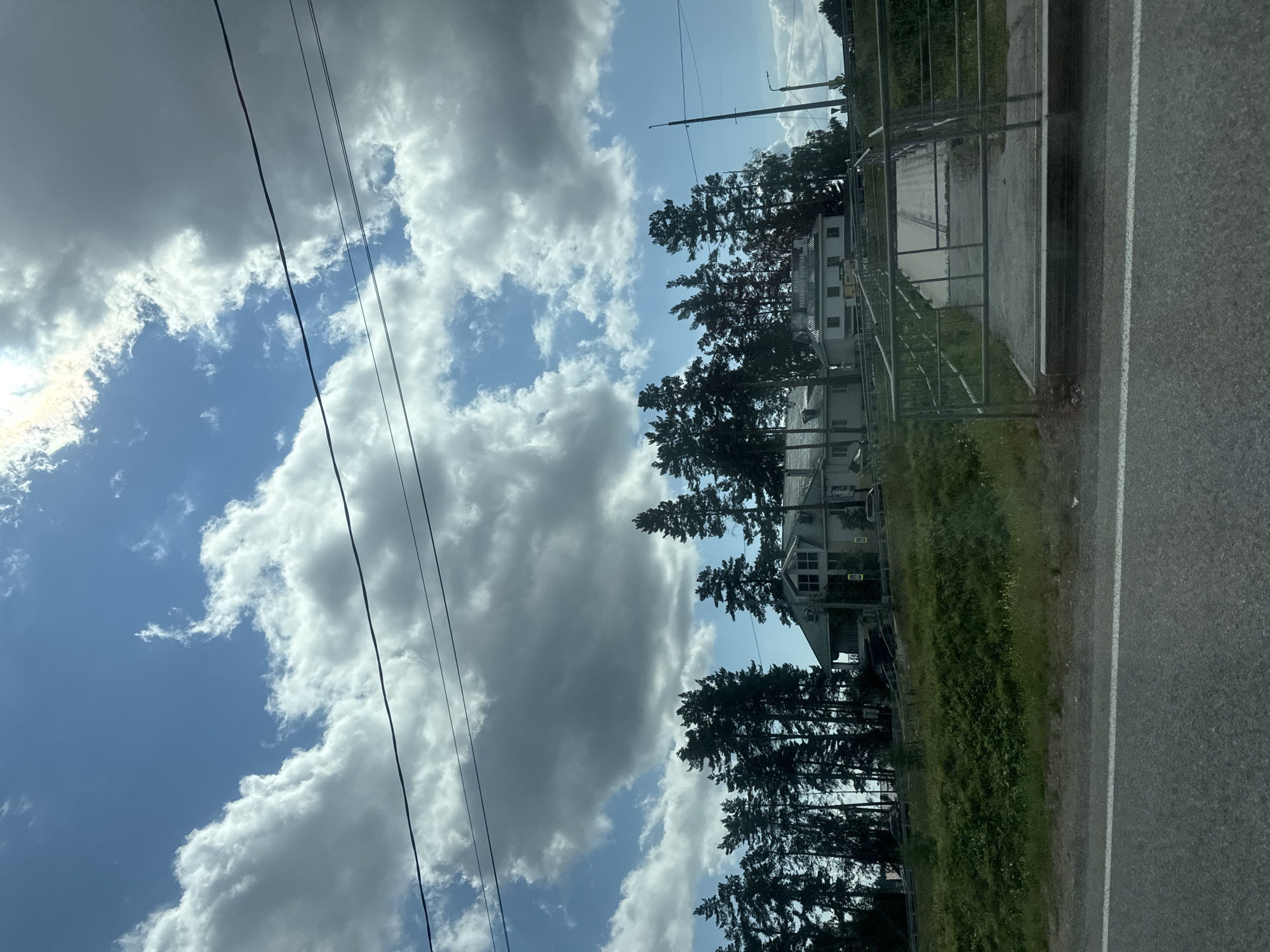 A slightly elevated, exterior, eye-level, horizontal, medium shot captures a rural scene dominated by a bright, partly cloudy sky and a line of tall evergreen trees. In the mid-ground, several light-colored buildings with dark roofs are nestled among the trees, one with a distinctive multi-pane window structure. To the right, a pedestrian bridge with metal railings crosses a grassy area, leading towards the buildings. The foreground features a paved road with a white line and a grassy embankment. Power lines are visible stretching across the sky. The image is taken from a moving vehicle, as evidenced by the slight motion blur on the road and embankment. The location is specified as Novolisiha, Russia. There are no people visible in the image.
