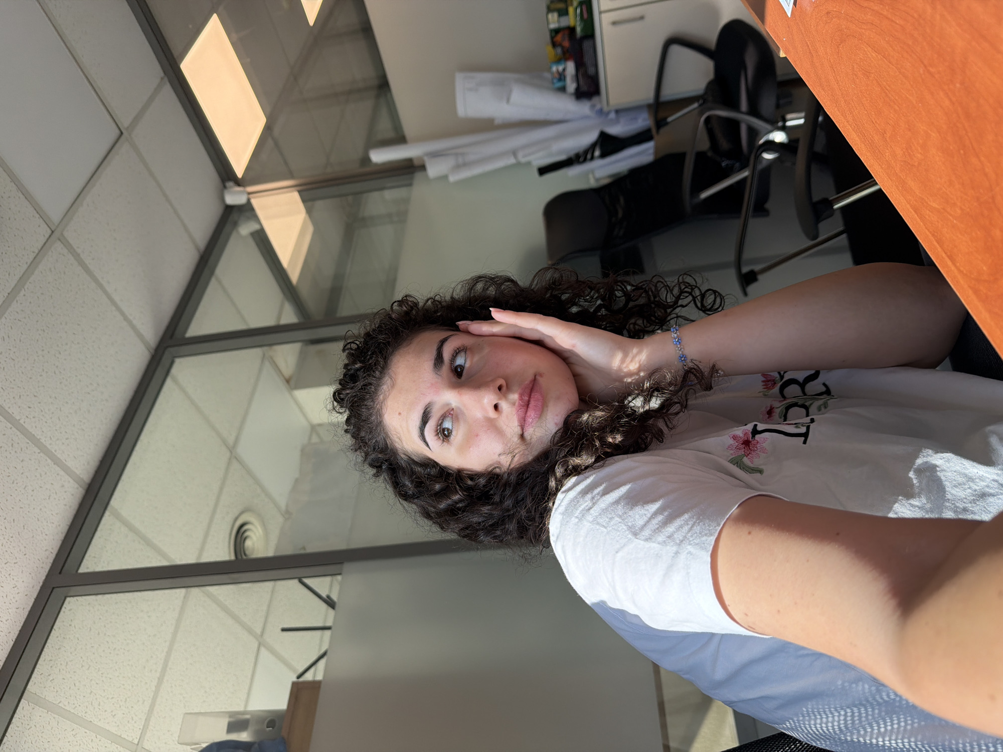 The image depicts a young woman with dark curly hair, wearing a white t-shirt with floral embroidery. She is indoors, in what appears to be an office environment, with large glass partitions and a suspended ceiling with fluorescent lighting. The woman is seated, with her left arm resting on a wooden desk that is orange in color. She is holding her right hand to her cheek, looking upwards and to her left, with a contemplative expression. In the background, there are office chairs, rolled-up documents, and a cabinet with various items on it. The lighting suggests it is daytime, with sunlight casting shadows. The setting does not provide any specific indicators to determine the location as Sofia, Bulgaria.