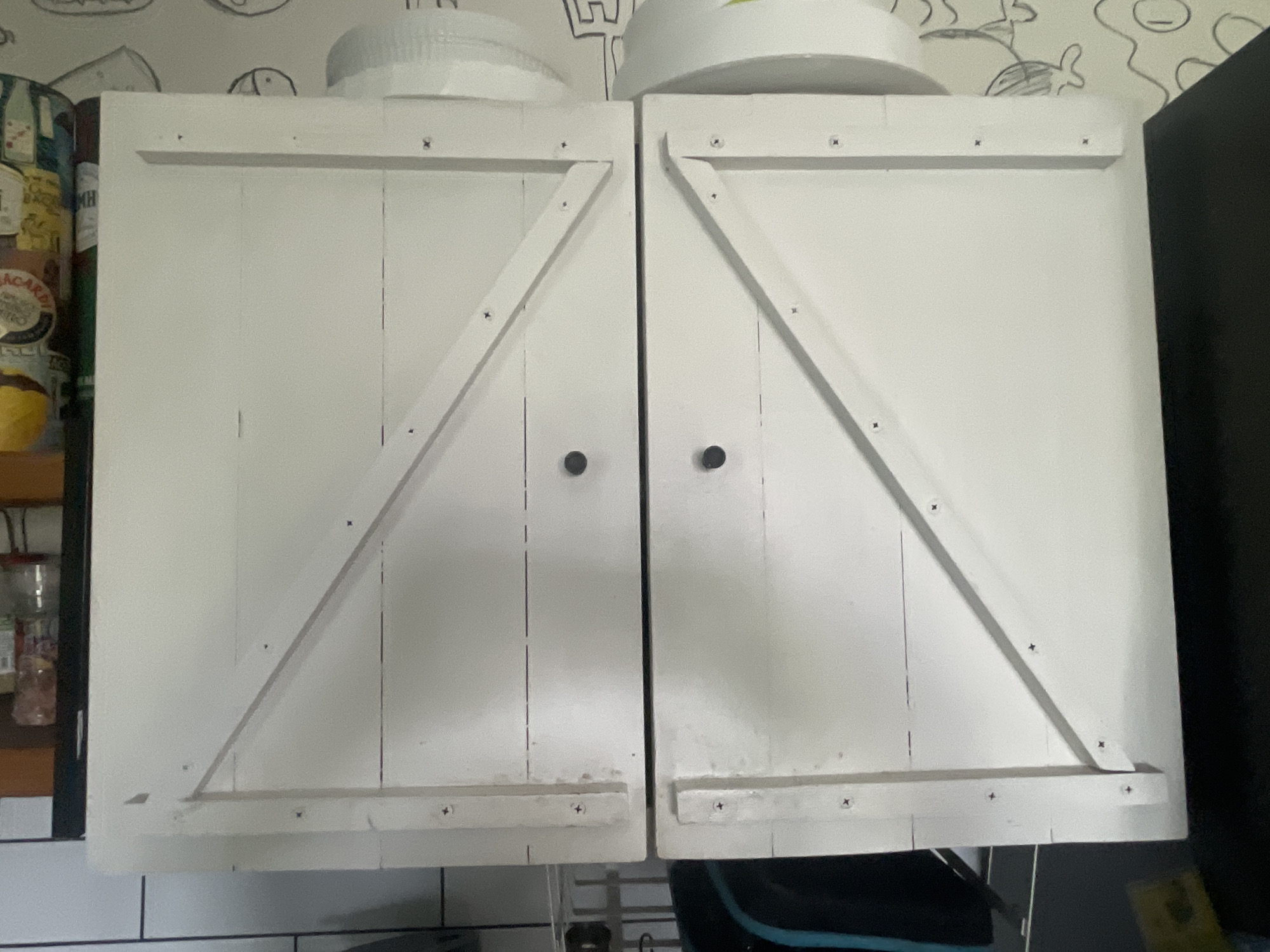 In the city of Мухавецкі сельскі Савет, Belarus, the image displays a close-up view of two white cabinet doors with a decorative "X" design on each. The doors have visible screws and appear to be made of wood. There are two dark circular knobs on each door. To the left of the cabinets, a shelf with bottles of alcohol is partially visible. The background features a wall with a patterned wallpaper and a dark object to the right. The cabinets are positioned above a tiled surface.