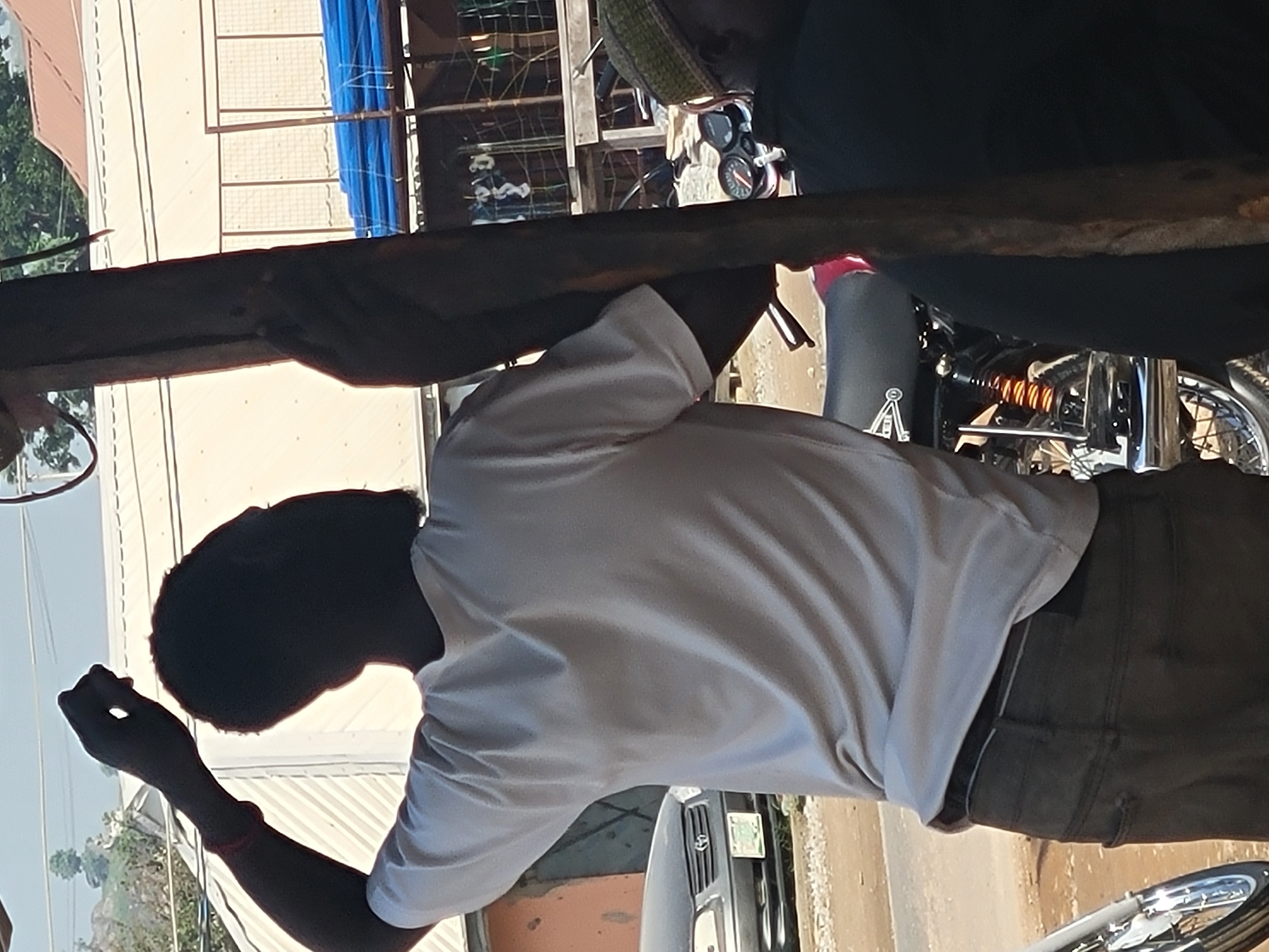 In Suleja, Nigeria, a man wearing a light-colored t-shirt and dark pants stands with his back to the camera, his right arm raised and hand clenched. He appears to be holding onto a wooden pole. Beside him, a motorcycle is visible, with another person partially visible in the foreground wearing a green cap and dark clothing. In the background, a dark-colored car is parked on a dirt road, with buildings and blue tarpaulin structures further beyond. The overall scene suggests an outdoor setting possibly in a market or along a street.
