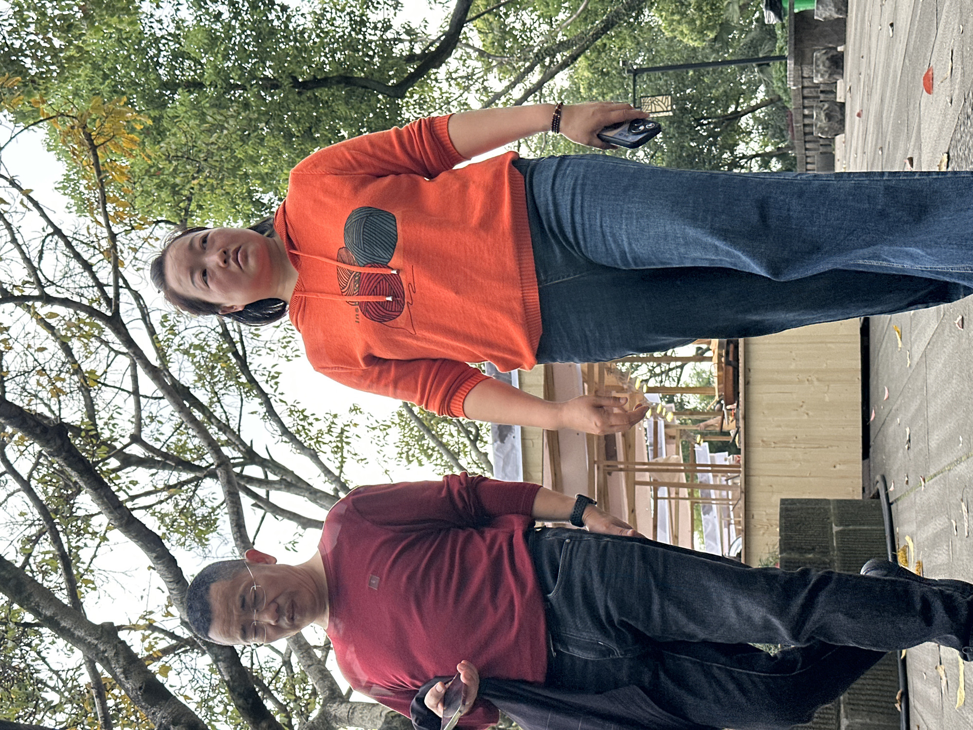 Two people, a male and a female, stand outdoors on a paved surface, possibly a sidewalk or plaza. The male figure, closer to the camera, wears glasses, a long-sleeved red shirt, and dark pants. He holds a dark object in his left hand. The female figure wears an orange hooded sweatshirt with a graphic on the front, blue jeans, and holds a cell phone in her right hand. The background includes bare tree branches and green foliage, suggesting a park or urban green space. There is a glimpse of a wooden structure and a section of wall. Given the prompt, the location is likely within Yuzhong District, China.
