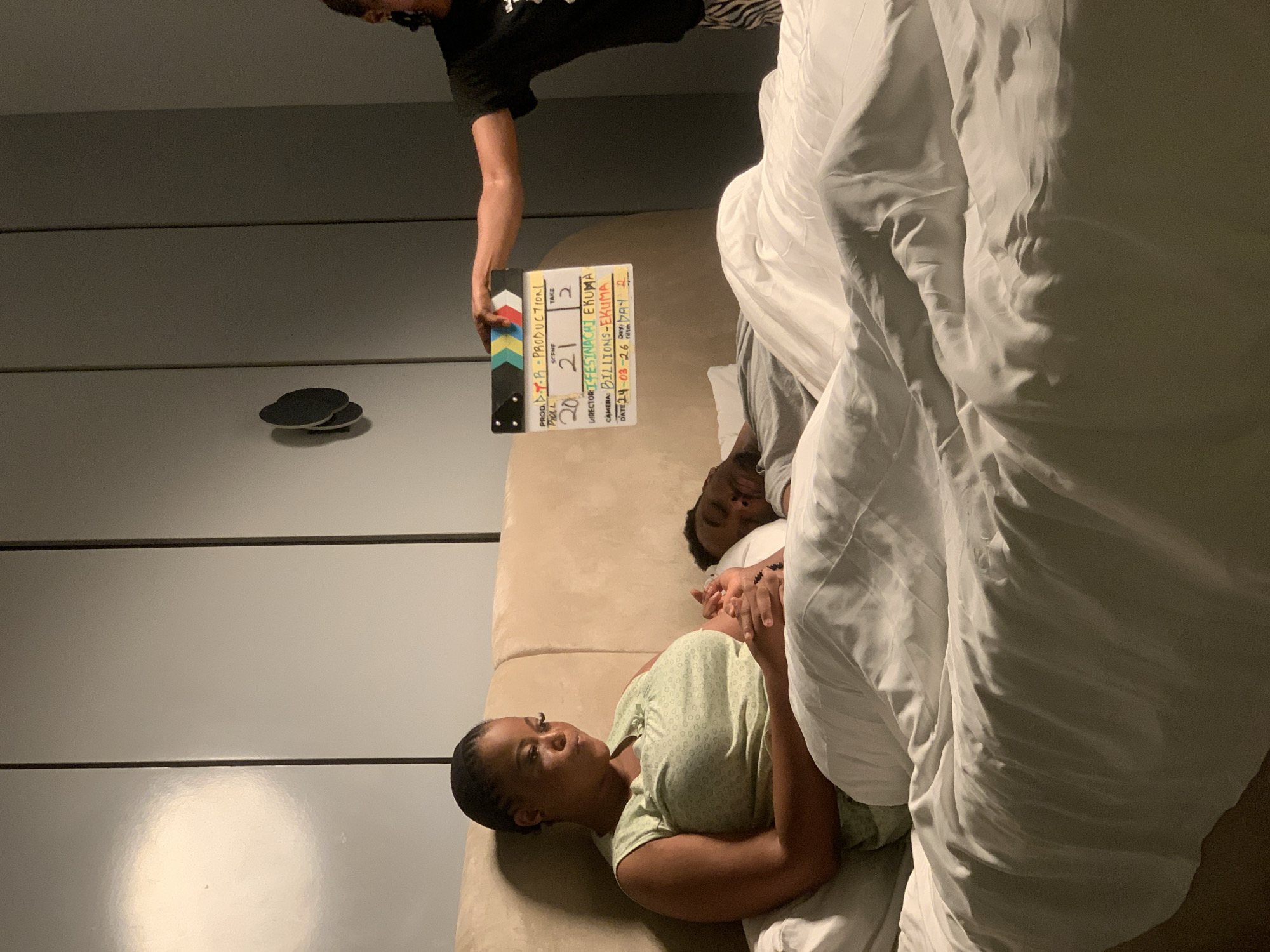 The image displays an indoor scene, likely a bedroom, functioning as a film set. In the foreground, a dark-skinned woman with dark hair is seated upright in a bed with a light brown upholstered headboard. She wears a light green sleeveless top and holds the hand of a dark-skinned man lying beside her, who appears to be asleep or unconscious, with his eyes closed. White bedding covers both individuals from the waist down. To the right, a third dark-skinned individual, partially visible and wearing a black t-shirt and patterned pants, holds an open clapperboard above the bed. The clapperboard reads "PROD: T.R PRODUCTION," "ROLL: 20," "SCENE: 21," "TAKE: 2," "DIRECTOR: FESTINACHI EKUMA," "CAMERA: BILLIONS-EKUMA," and "DATE: 24-03-26 DAY: 2." The background consists of dark grey paneled walls, featuring a dark, circular wall decoration on the right and a distinct light reflection on the far left. The specific city location of Moba, Nigeria, is not discernible from the visual content of the image.