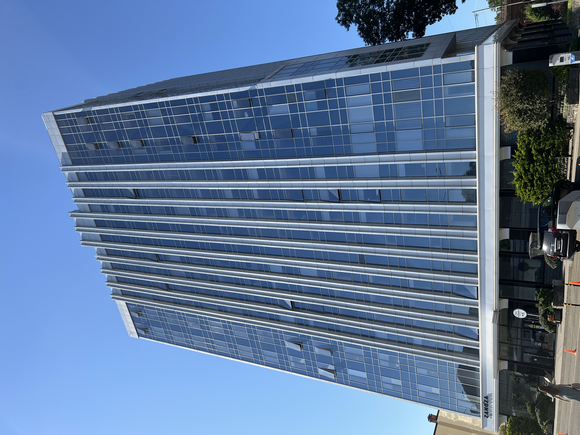 This is a low-angle, wide-shot photograph of a modern, multi-story office building with a glass facade and vertical metallic accents, set against a clear blue sky. The building features a prominent sign at its base that reads "ZANOZA TO GO". Several windows are open on various floors of the building. In the foreground, on the sidewalk and street, there are some parked vehicles, including a black SUV with a license plate, orange traffic cones, and some landscaping with bushes and trees. A woman wearing a light-colored dress is walking away from the camera on the sidewalk. The building is located in the city of Krasnodar, Russia.