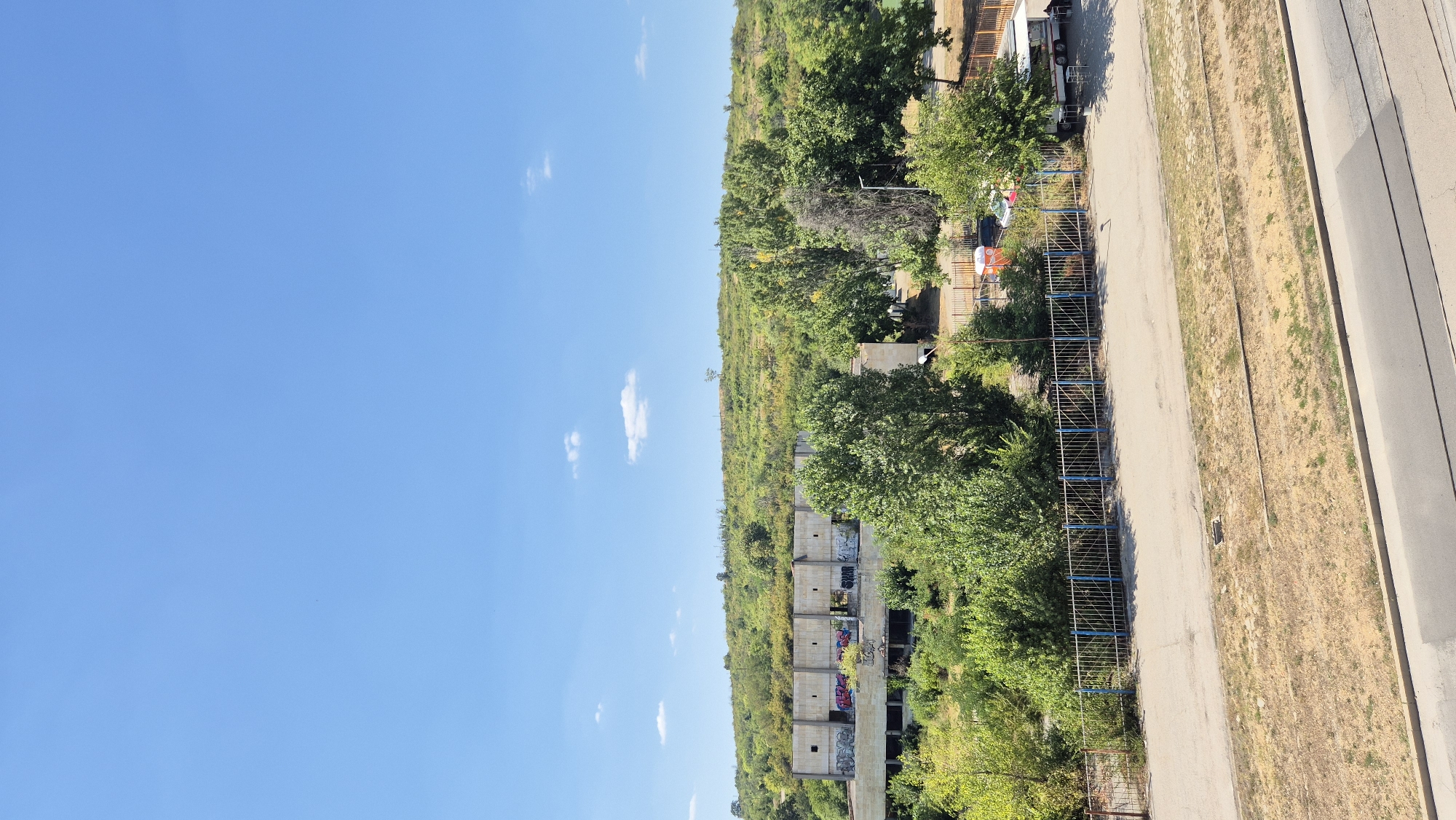 This is a vertical, eye-level, outdoor photograph taken on a clear, sunny day in Lovech, Bulgaria. The image captures a view of an abandoned concrete building covered in graffiti, situated amidst overgrown vegetation and trees. In the foreground, a paved road runs horizontally, with a dry, grassy verge bordering it. A blue-railed fence separates the road from a grassy area with trees and the dilapidated building. Further in the distance, a tree-covered hill forms the backdrop under a bright blue sky with a few scattered white clouds. A small white trailer is parked near the fence in the middle ground.