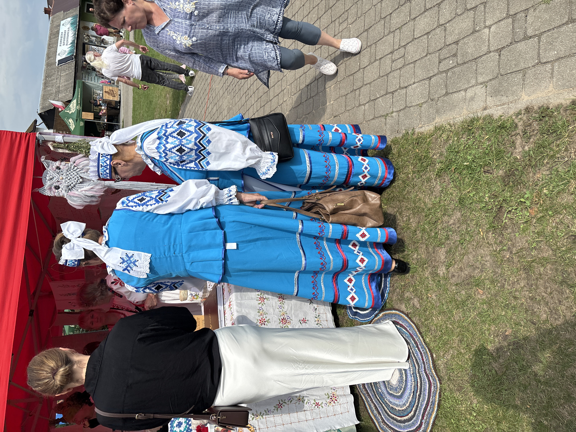 In the city of Podlewkowie, Poland, at a local event, two women dressed in traditional blue and white embroidered folk costumes stand near a red tent. The costumes feature intricate geometric patterns on the sleeves and skirts, with white headpieces adorned with large white bows. One woman wears a black shirt and a long, flowing white skirt, while the other has a white shirt with embroidered blue details. They are positioned in front of a table covered with a white tablecloth featuring floral embroidery. A woven circular mat is visible on the ground near their feet. In the background, other people are browsing stalls, and a building with a gray roof is visible, bearing a sign that appears to be in Polish. The ground is a mix of grass and a paved walkway.