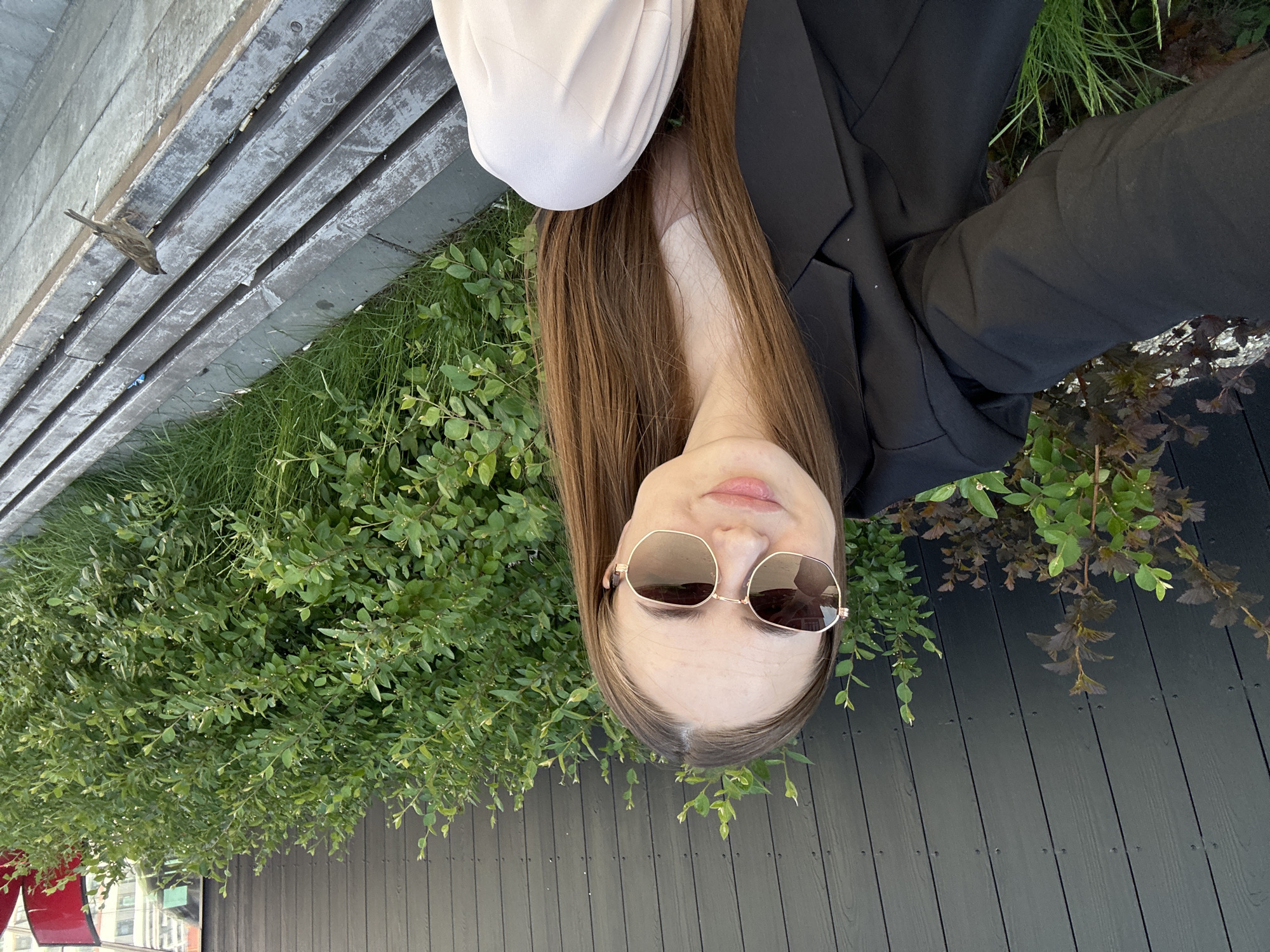 A woman with long brown hair, wearing sunglasses, a black blazer, and a white mask, is taking a selfie outdoors. She is positioned near a wooden structure with greenery growing around it. A bird is perched on the wooden structure. The environment suggests an urban setting. There is no location information.