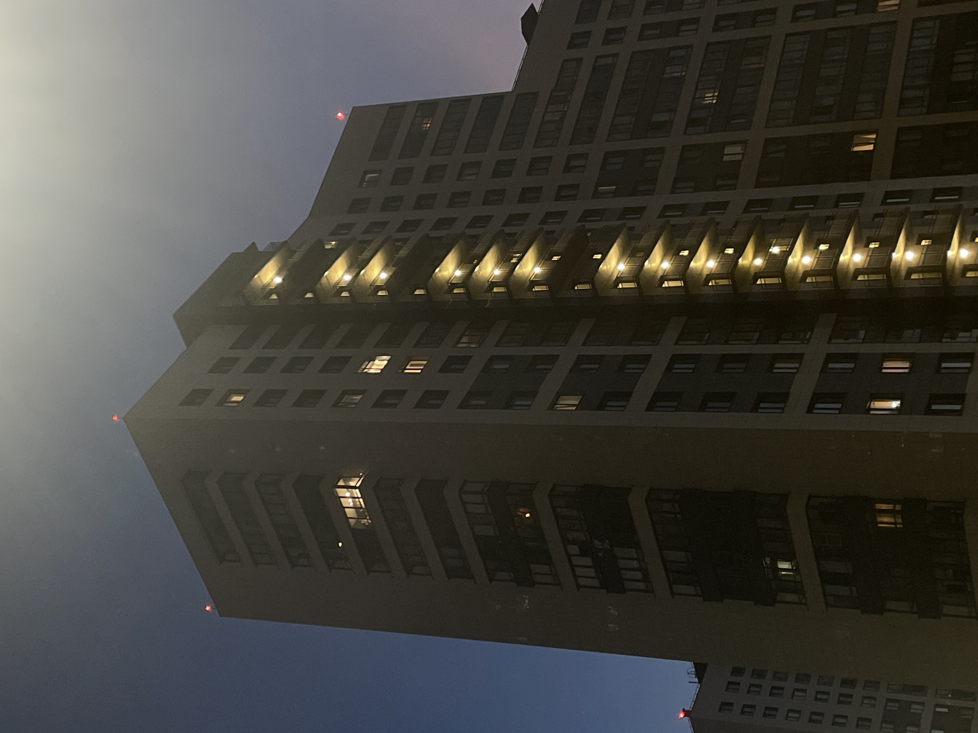 The image shows a tall, modern apartment building in Sinyavino, Russia, at dusk or dawn. The building is mostly dark, with some windows illuminated from within, suggesting residents are home. The sky is a deep blue, with a hazy quality, perhaps due to fog or mist, obscuring the upper portions of the building and the surrounding environment. Red warning lights are visible at the top of the building, indicating its height. The architecture features repetitive window patterns and recessed balconies with integrated lighting, which casts a warm glow on the facade. Another building, partially visible at the bottom of the frame, suggests a dense urban or suburban setting. The overall mood is quiet and somewhat mysterious, characteristic of twilight in a city. There is no visible text in the image.