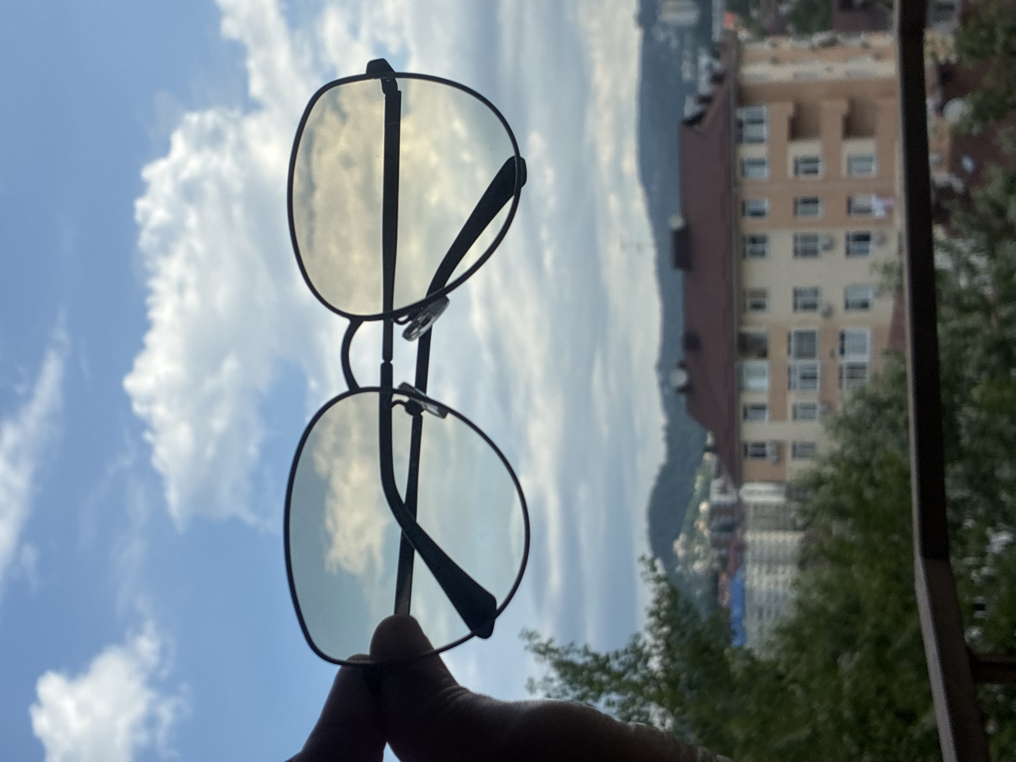 A close-up, handheld shot features a pair of black-rimmed sunglasses held up against a bright blue sky with scattered white clouds. The lenses of the sunglasses reflect the sky and clouds. In the background, out of focus, is a city skyline with several multi-story buildings. One prominent building is a tan and beige apartment complex with a dark red roof, featuring numerous windows and balconies. Dense green trees and foliage are visible in the foreground and mid-ground, obscuring parts of the city. A dark brown railing runs across the bottom of the frame. The image appears to be taken from a balcony or elevated vantage point, overlooking a cityscape with hilly terrain in the distance. The setting is suggestive of a sunny day in Sochi, Russia, given the architectural style and the presence of greenery.