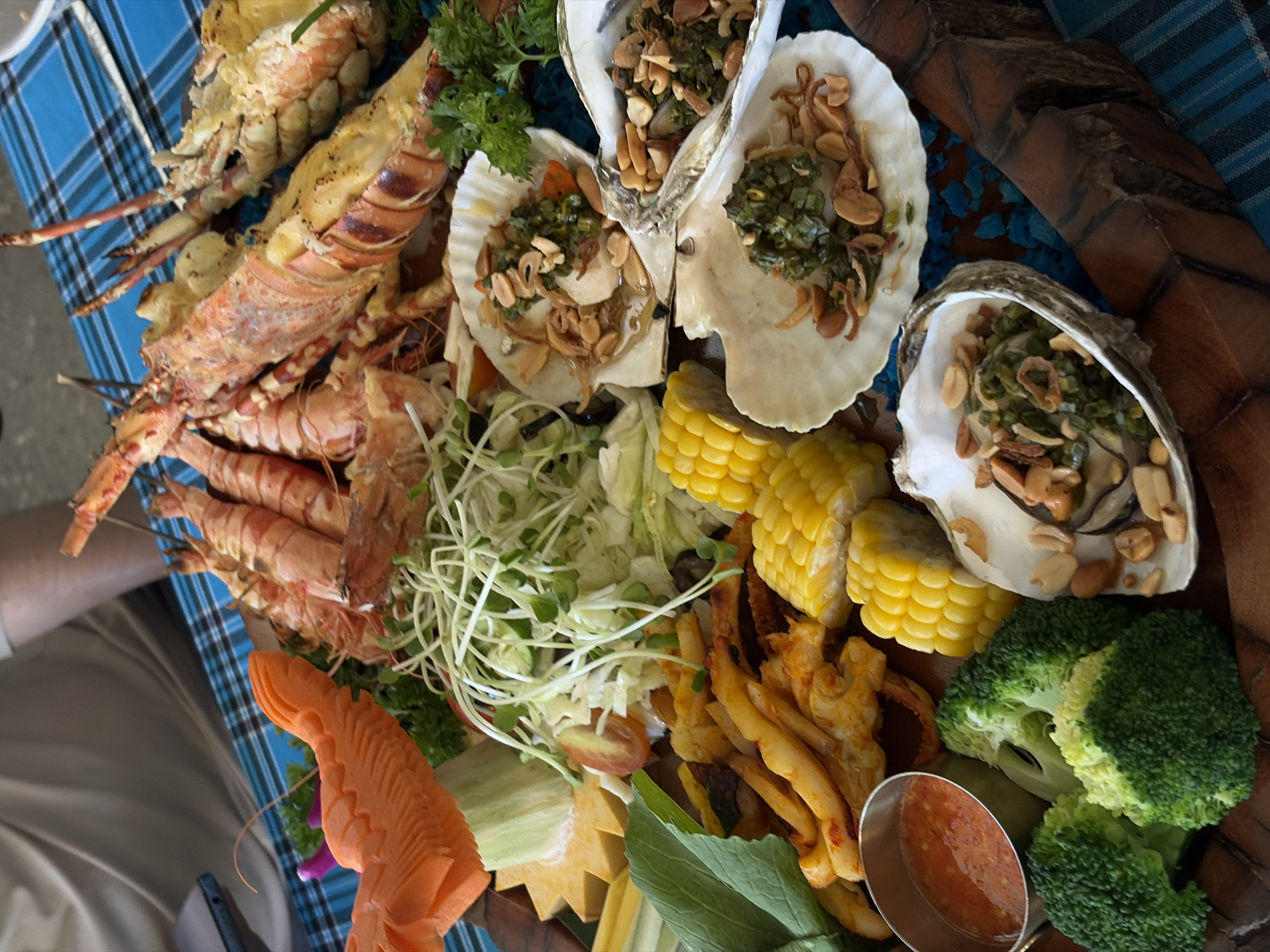 The image captures a generous seafood and vegetable platter, suggesting a feast in a coastal setting like Đà Nẵng, Vietnam. The large, rustic dark brown serving tray is laden with various cooked delicacies.
Dominating the platter are several large lobsters, some appearing split open and topped with a golden, possibly buttery or cheesy, mixture. Alongside them are numerous cooked prawns or shrimp, neatly arranged on skewers. Multiple oysters and scallops, still in their shells, are visible, each topped with a vibrant mixture of chopped green herbs (likely scallions), fried garlic, and roasted peanuts, a common preparation in Vietnamese cuisine.
The seafood is accompanied by an assortment of fresh vegetables and sides. These include bright yellow corn on the cob cut into rounds, and crisp green broccoli florets. A fresh salad is also present, featuring shredded lettuce, white sprouts, and what appears to be sliced cucumber and cherry tomato halves. Additionally, there are golden-fried calamari or similar seafood sticks. The presentation is enhanced by decorative garnishes such as fresh parsley, slices of star fruit, other leafy greens, and an intricately carved orange carrot, possibly depicting a fish or dragon. A small metallic cup holds a reddish-orange, chunky dipping sauce.
A person's arm and lower body, wearing khaki pants, are partially visible on the left, indicating someone is seated at a table. A blue and black plaid tablecloth is visible beneath the platter on this side. No specific time of day, weather, or visible text can be determined from the image, but the elaborate spread suggests a convivial dining event.