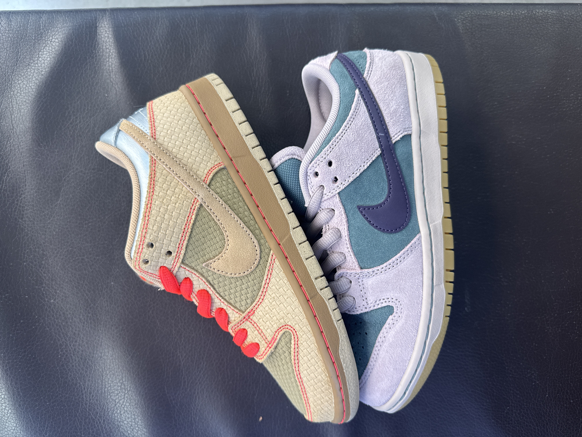 The image shows two Nike Dunk Low sneakers placed on a dark, textured surface. The sneaker on the left has a woven beige and olive green upper with a brown swoosh logo and red laces. The sneaker on the right has a blue and lilac suede upper with a purple swoosh logo and light gray laces. The location is not discernible from the image. There are no people in the image.