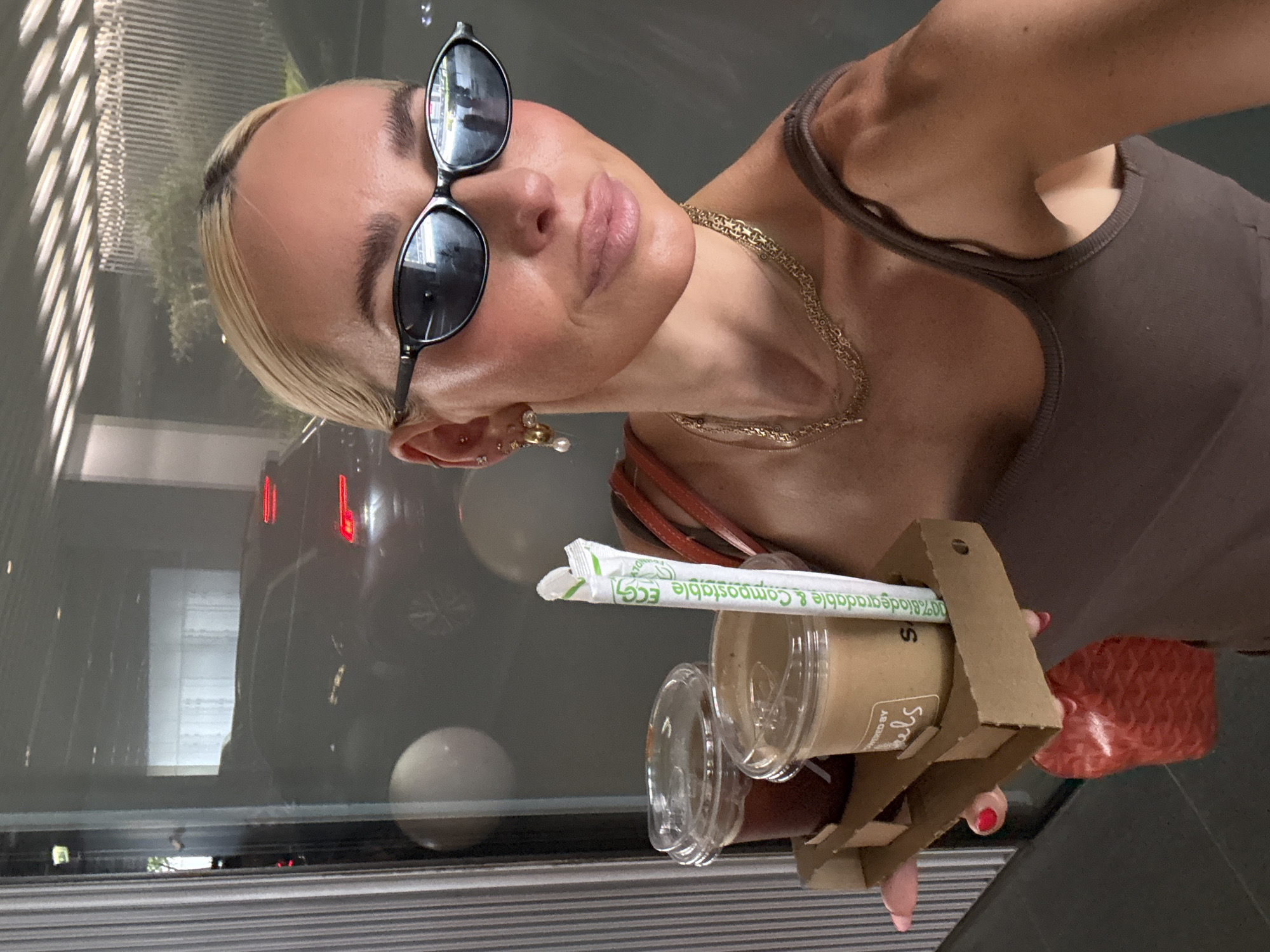 A woman with blonde hair pulled back, wearing dark sunglasses, a brown tank top, and gold jewelry, is taking a selfie in a modern, indoor setting. She is holding a cardboard drink carrier with two iced coffee beverages and a compostable straw. A red patterned bag hangs from the carrier. Reflections in the large glass windows behind her show a street scene with a dark car. The smooth, grey floor suggests a polished concrete or similar material. The interior lighting is bright, with linear fixtures visible on the ceiling, indicating a commercial or public space. The overall ambiance is clean and contemporary, possibly a hotel lobby or a high-end retail environment.